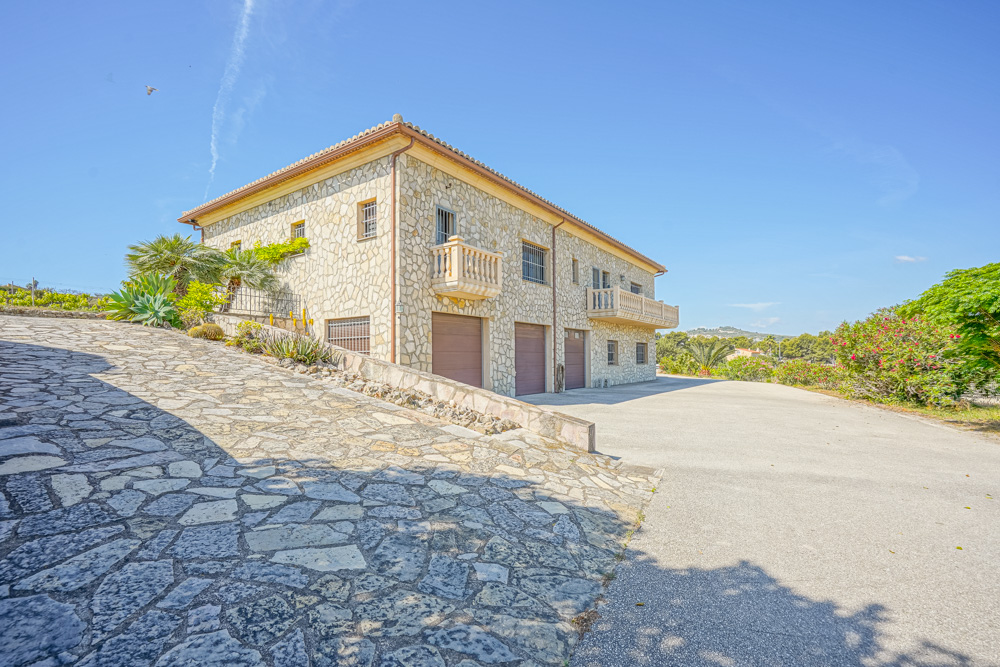 Fachada de piedra natural con garaje triple, rampa de acceso empedrada y balcones clásicos rodeados de vegetación mediterránea y cielo despejado.