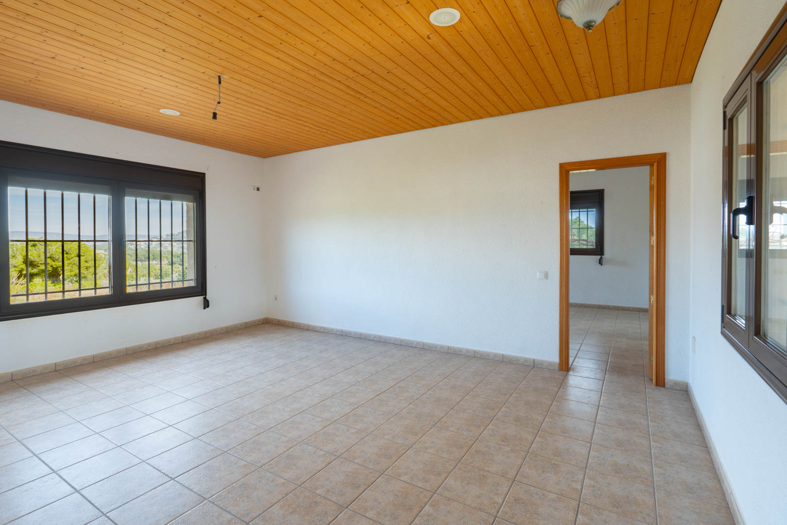 Habitación con techo de madera, suelos de gres beige y ventanas con marcos oscuros y rejas de seguridad que ofrecen vistas al entorno natural.