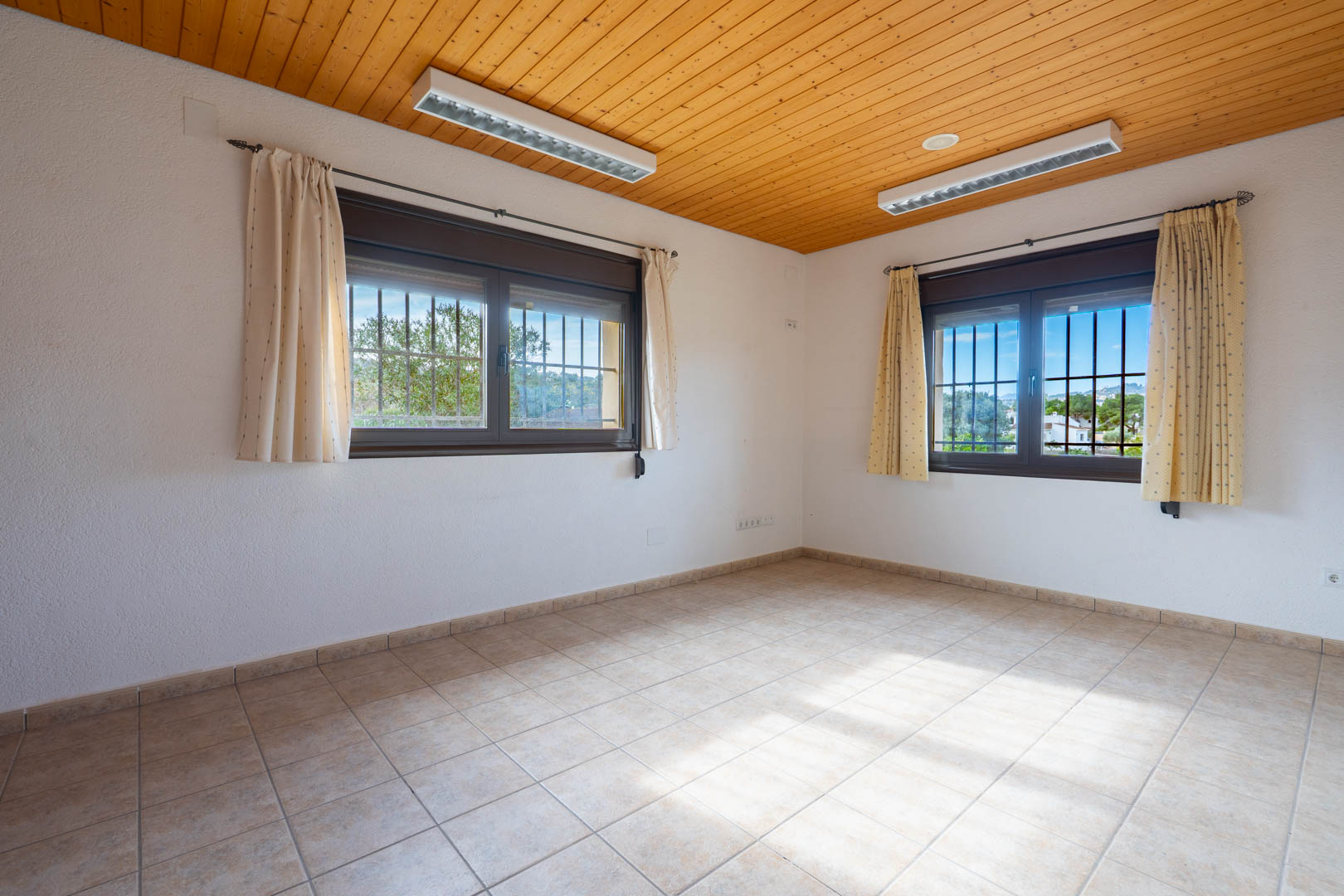 Habitación vacía con techo de paneles de madera natural, suelo de gres cerámico y ventanas dobles con rejas de seguridad y cortinas.