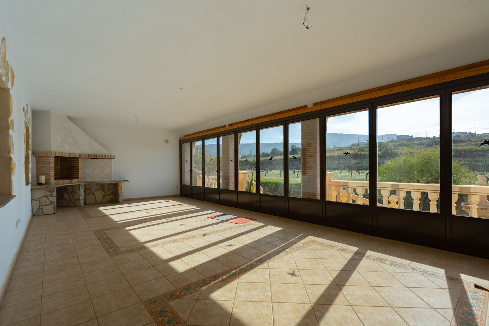 Terraza acristalada con barbacoa de piedra, suelos cerámicos con cenefas y amplios ventanales que ofrecen vistas despejadas al entorno natural.