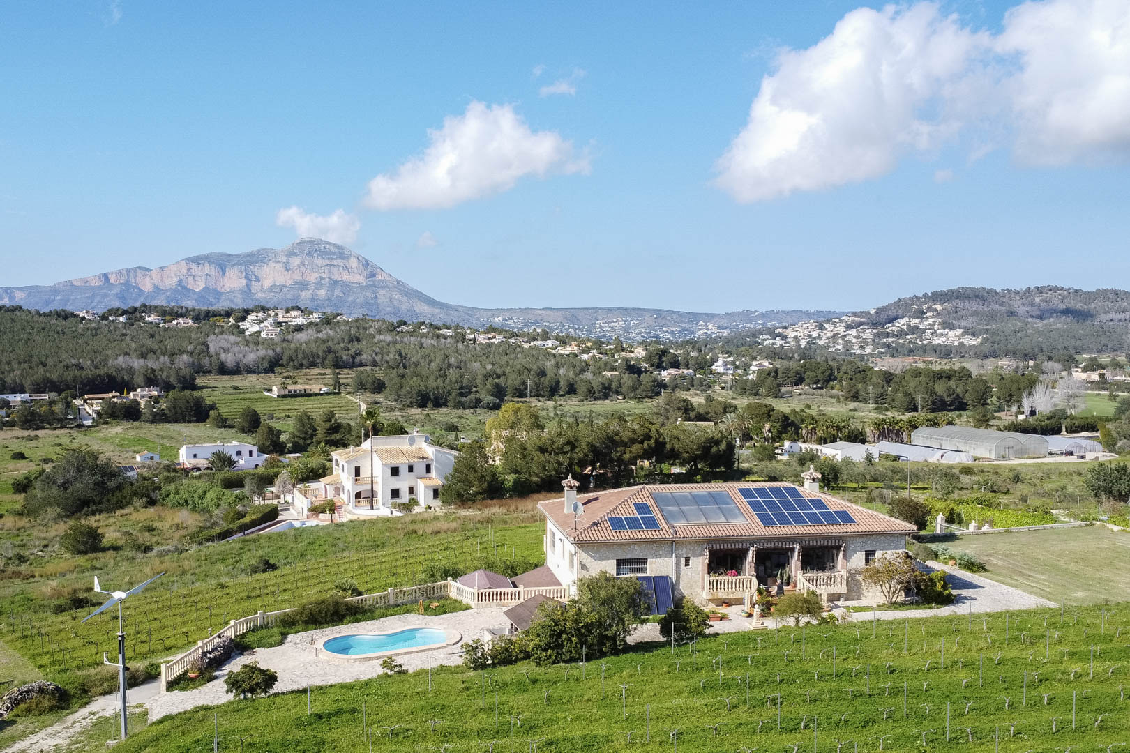 Vista aérea de una casa de piedra con paneles solares, aerogenerador y piscina, rodeada de campos de cultivo y con vistas panorámicas a la montaña.