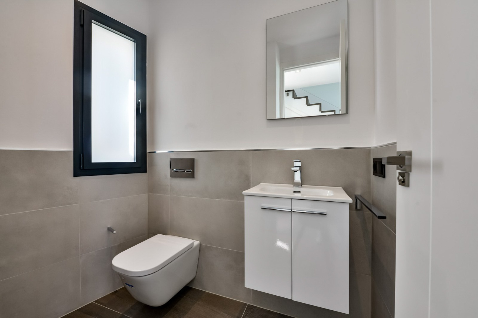 Aseo de invitados con inodoro suspendido, mueble de lavabo blanco de diseño minimalista y ventana de cristal traslúcido con marco oscuro.