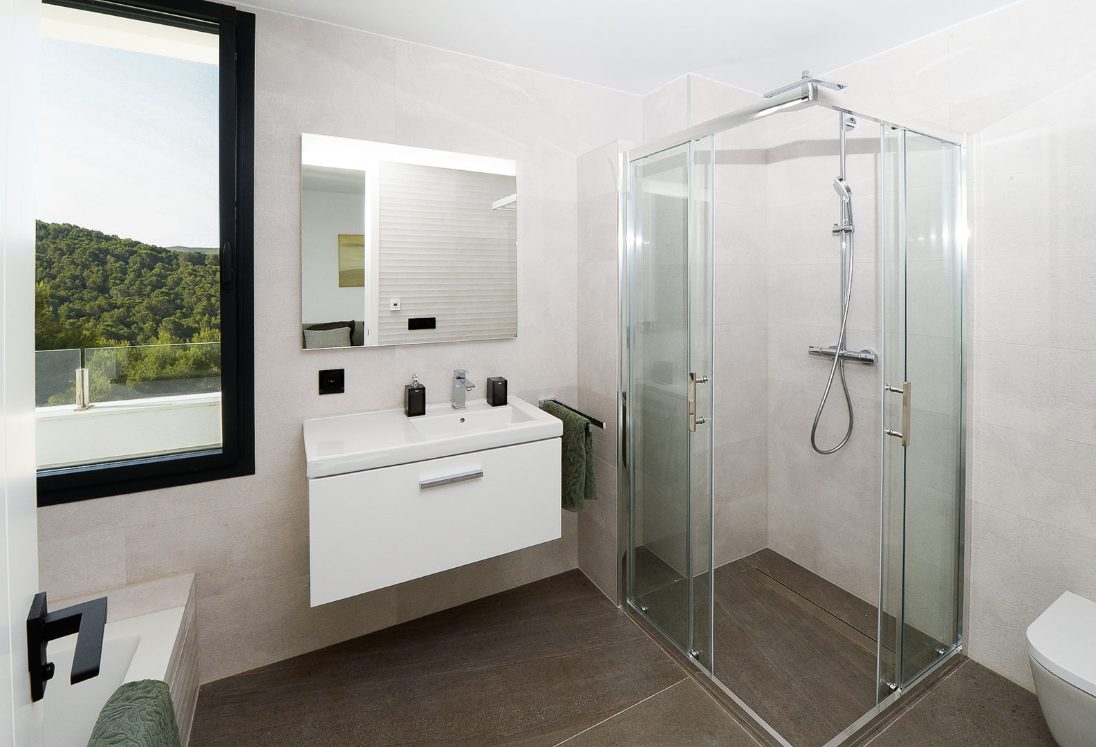 Baño moderno con ducha de cristal, mueble suspendido blanco y ventana con vistas a la montaña. Revestimientos en tonos neutros y suelo oscuro.