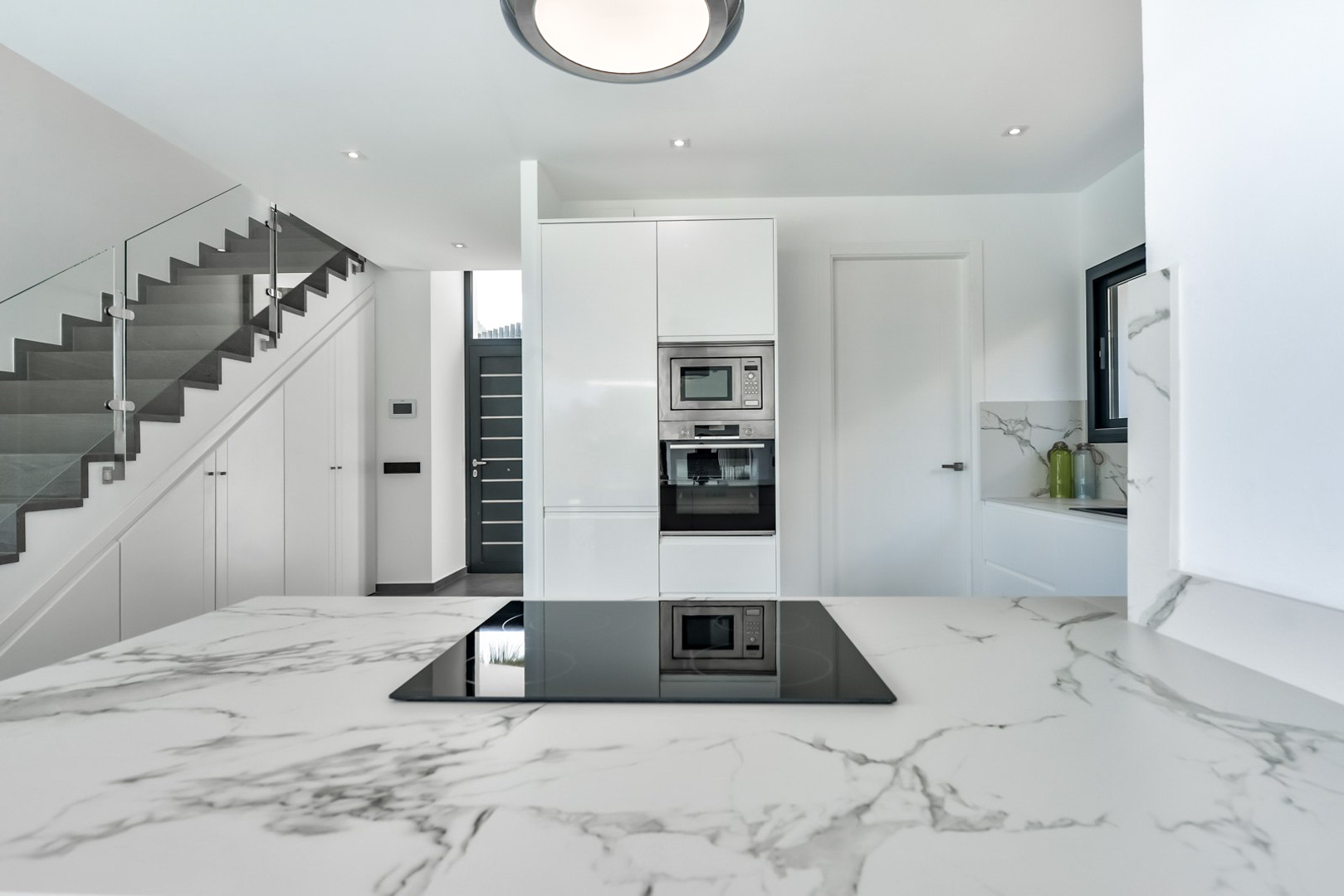 Cocina de diseño con encimera de mármol veteado y placa de inducción, junto a una escalera moderna con barandilla de cristal y armarios empotrados.