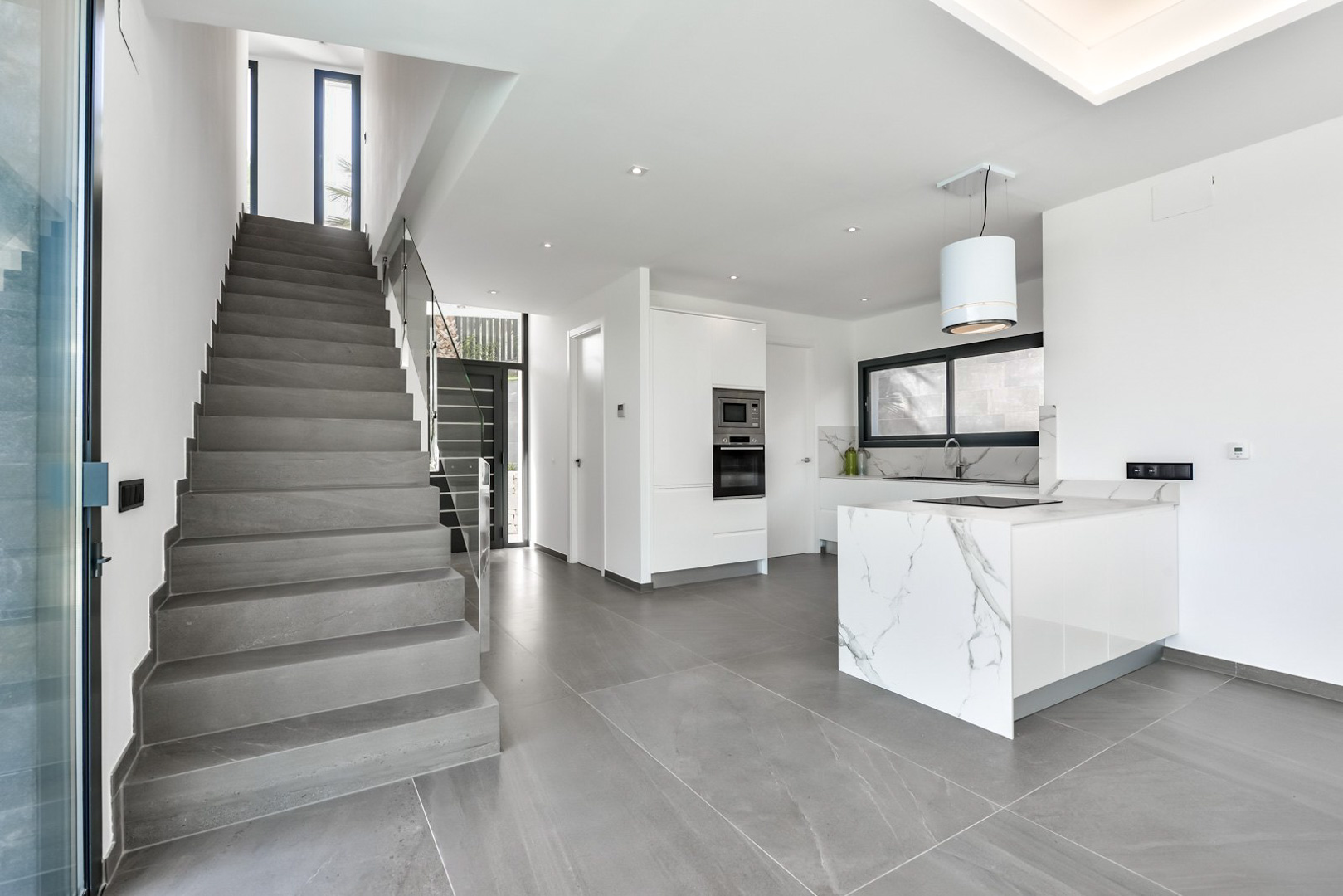 Cocina de diseño con isla de mármol y electrodomésticos integrados junto a una escalera de piedra gris con barandilla de cristal en un espacio diáfano.