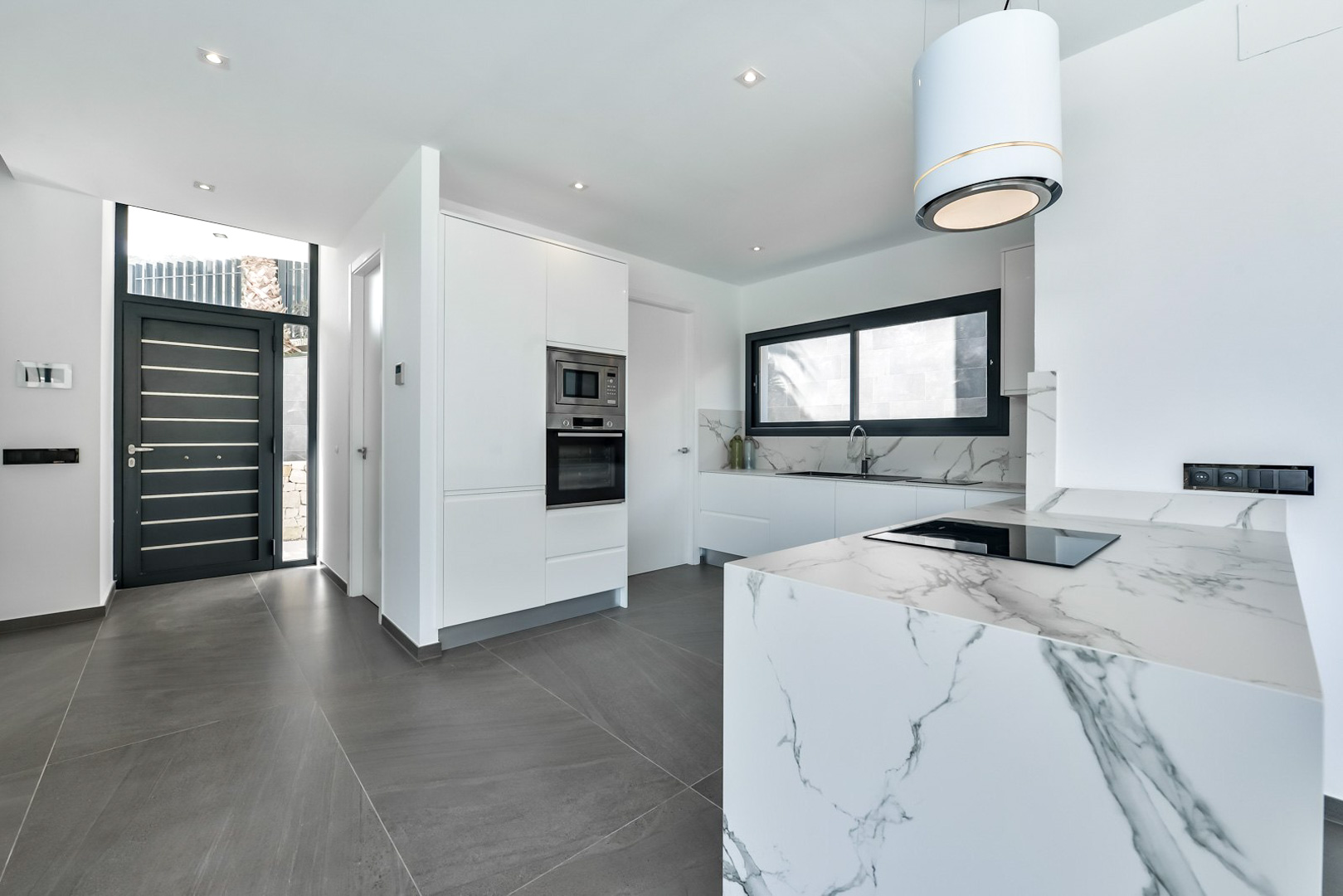Cocina moderna de diseño con isla de efecto mármol, electrodomésticos integrados y campana cilíndrica, situada junto a la entrada principal de carpintería negra.