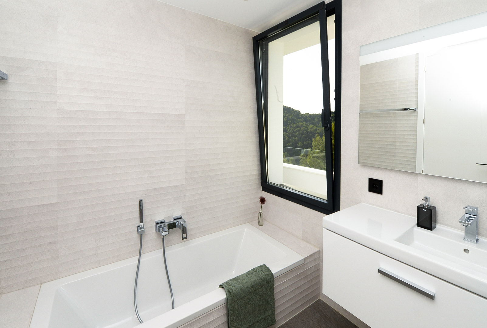 Cuarto de baño moderno con bañera encastrada, revestimiento cerámico texturizado y ventanal con vistas al entorno natural de Jávea.