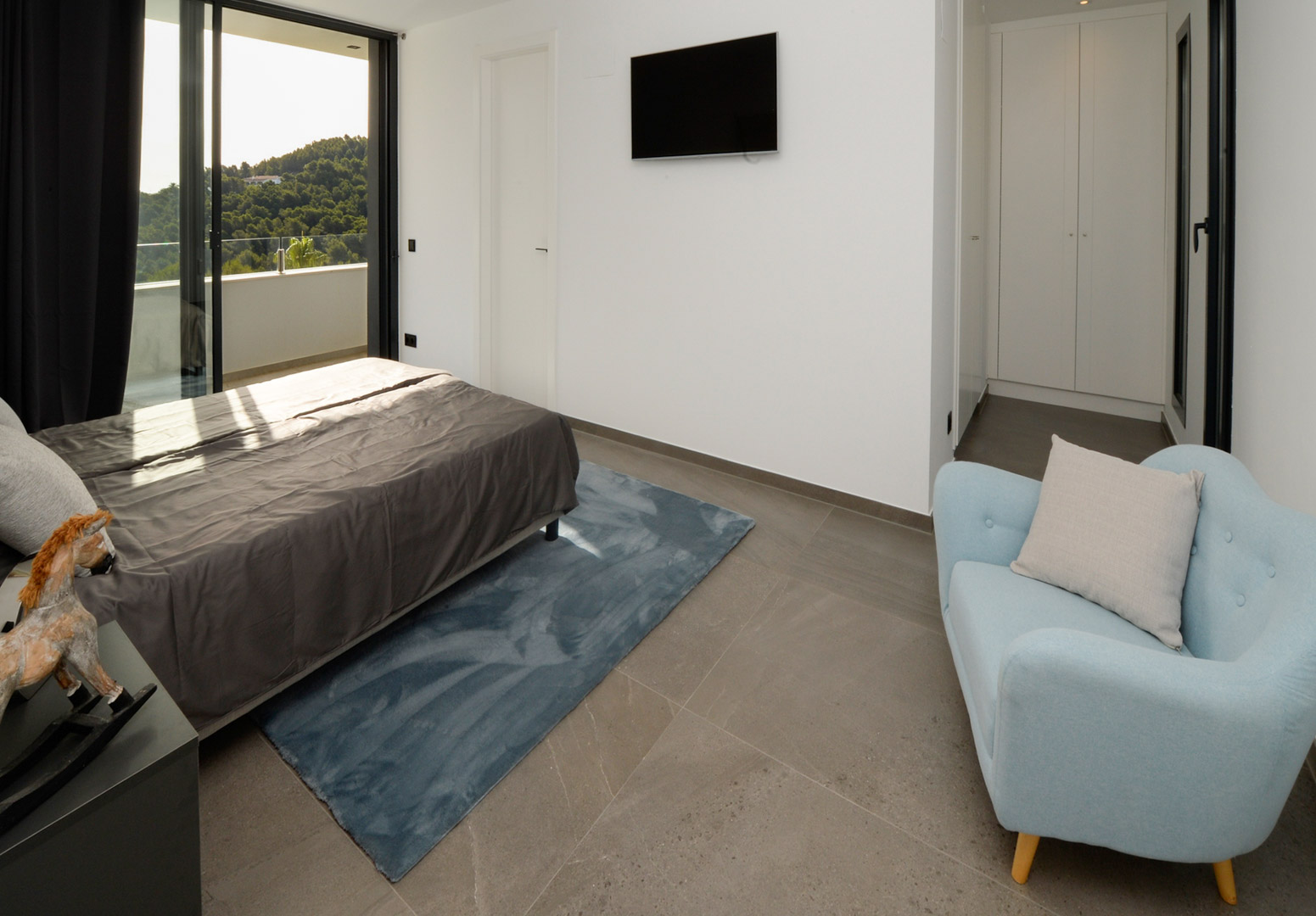 Dormitorio contemporáneo con acceso a terraza, suelos de gres gris, armarios empotrados blancos y vistas panorámicas a las colinas de Jávea.