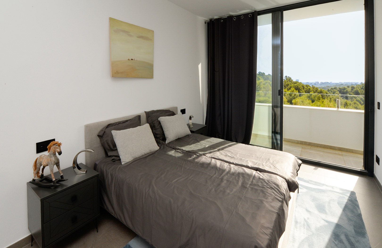 Dormitorio luminoso con cama doble en tonos grises, mesitas de noche modernas y gran ventanal corredero con acceso directo a la terraza y vistas al entorno natural.