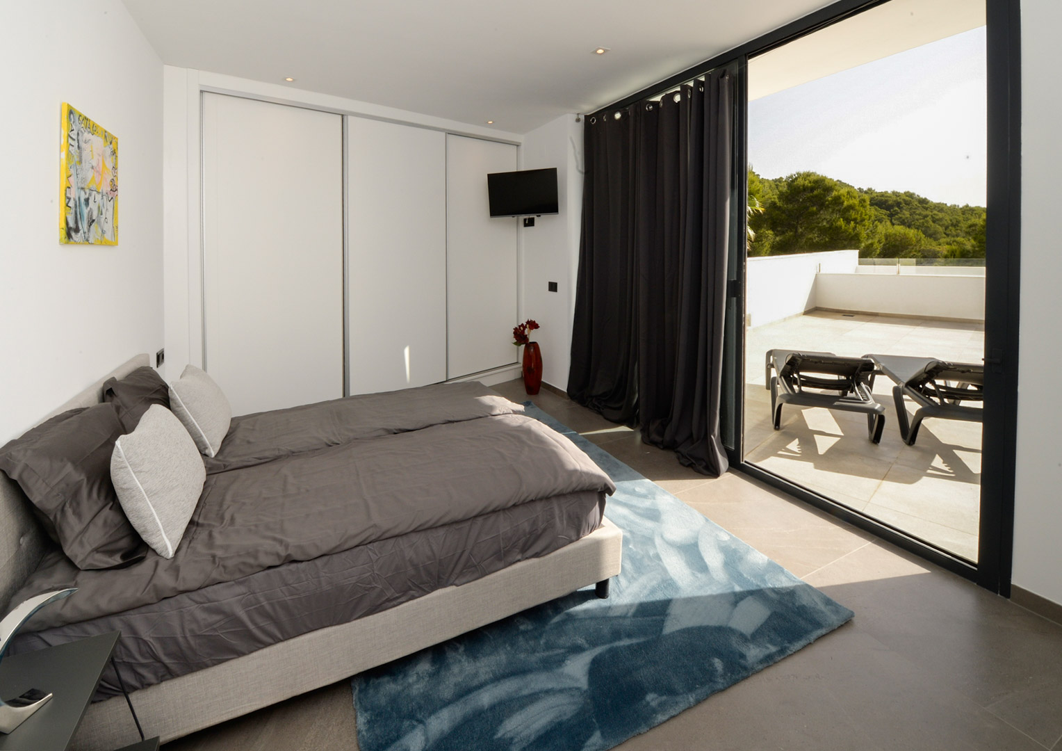 Dormitorio moderno con armarios empotrados blancos y acceso directo a una amplia terraza con tumbonas y vistas al entorno natural de Jávea.