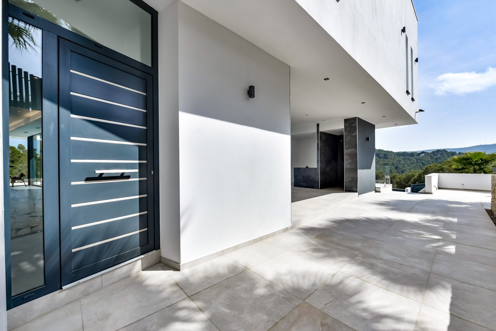 Entrada principal con puerta de diseño contemporáneo en azul oscuro y listones metálicos, porche de gres porcelánico y vistas despejadas al entorno natural de Jávea.
