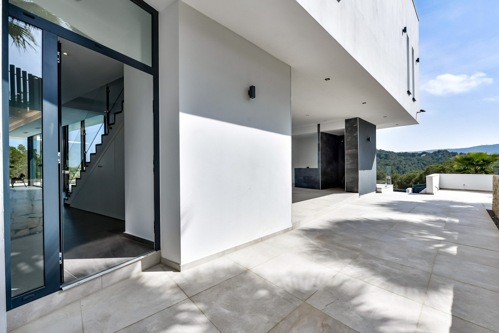Entrada principal y terraza cubierta con suelos de gres porcelánico, fachada blanca minimalista y vistas despejadas al valle de Jávea.