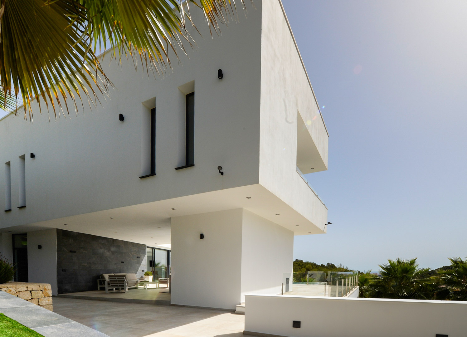 Fachada contemporánea con voladizo arquitectónico, paredes de piedra natural gris y terraza cubierta con mobiliario de exterior bajo iluminación integrada.