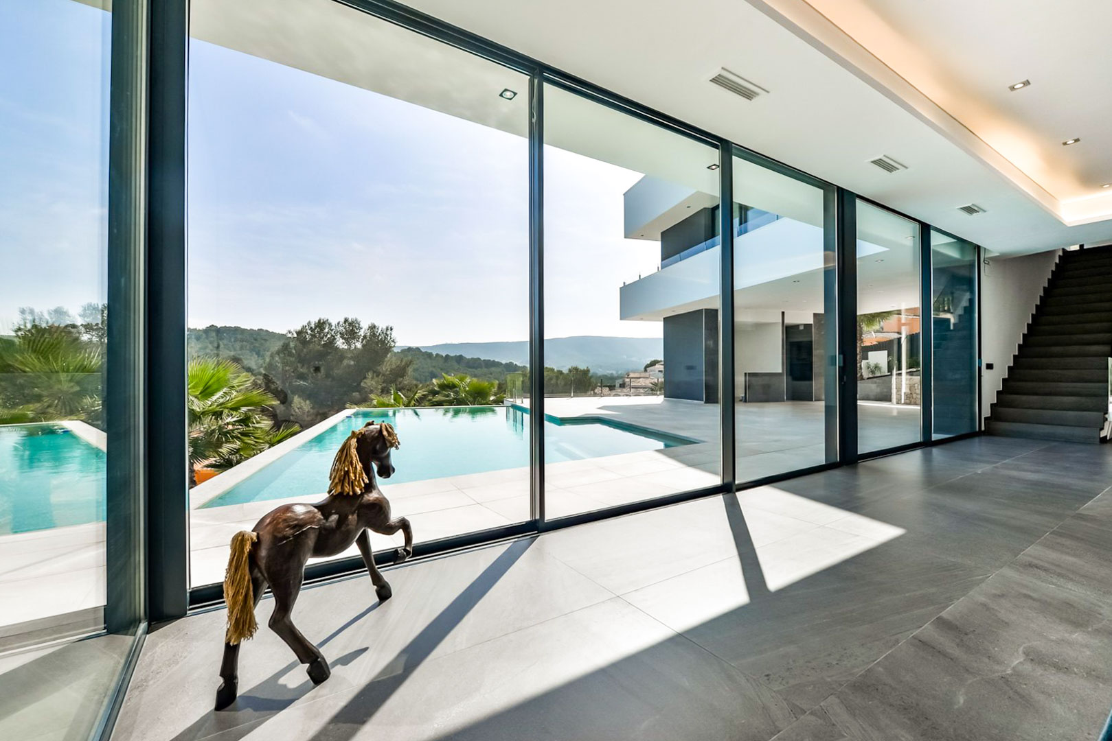 Salón con grandes ventanales de suelo a techo que integran la terraza y la piscina infinity. Suelos de gres cerámico gris y escalera de diseño con vistas al mar.