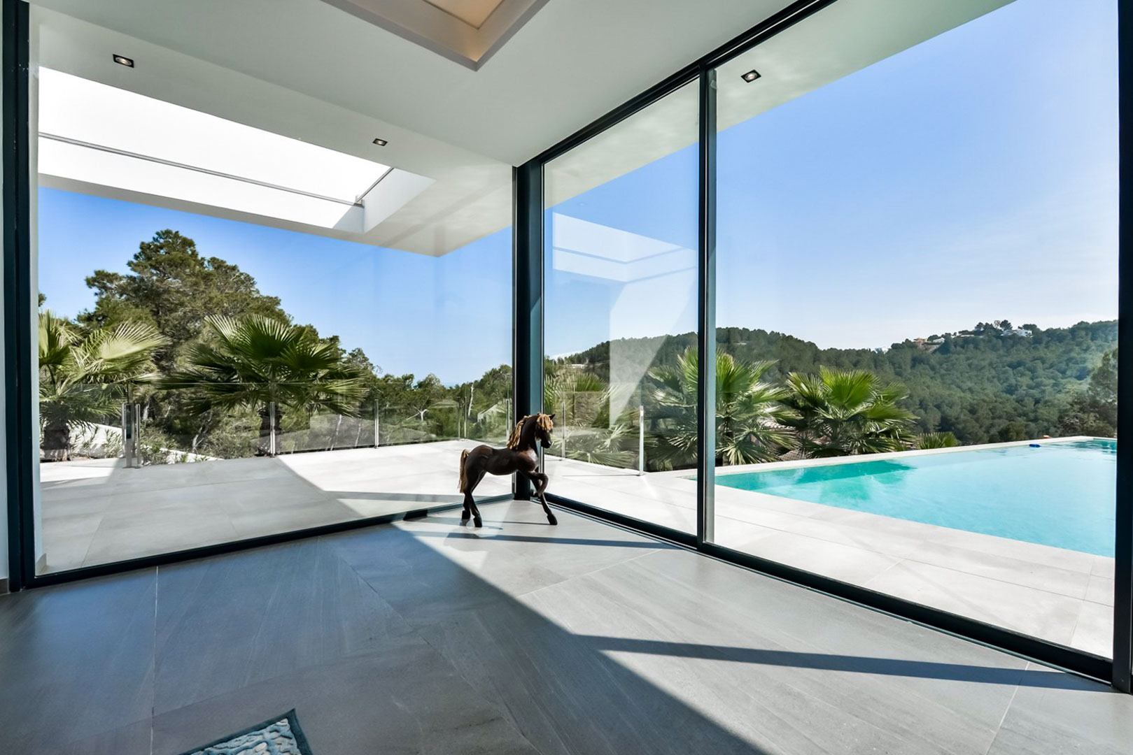 Salón con ventanales de suelo a techo que integran la terraza exterior, la piscina infinity y las vistas panorámicas a las colinas verdes de Jávea.