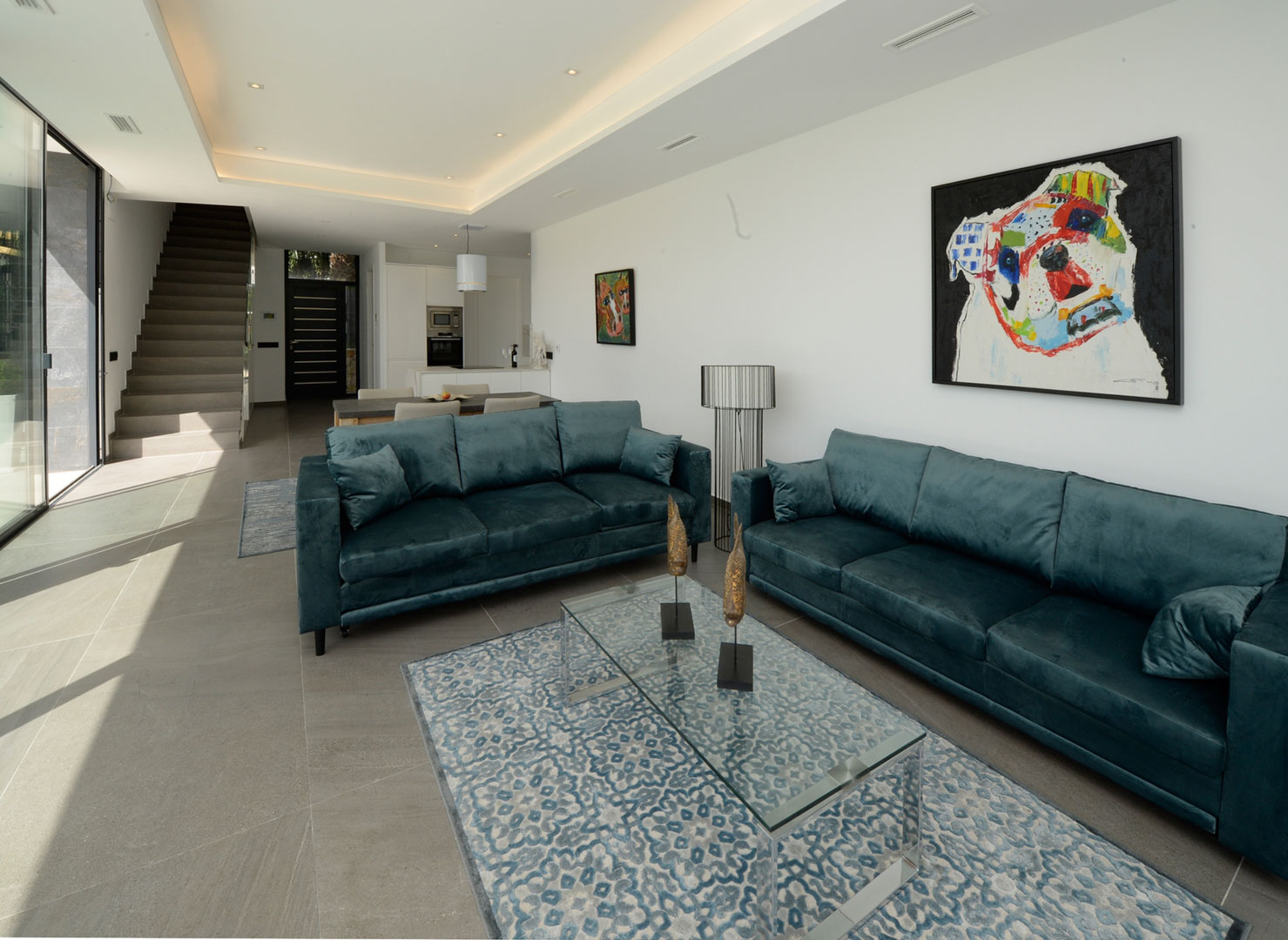 Salón de concepto abierto con sofás de terciopelo azul, mesa de cristal y suelos porcelánicos. Incluye iluminación LED empotrada y acceso directo a la escalera y cocina.