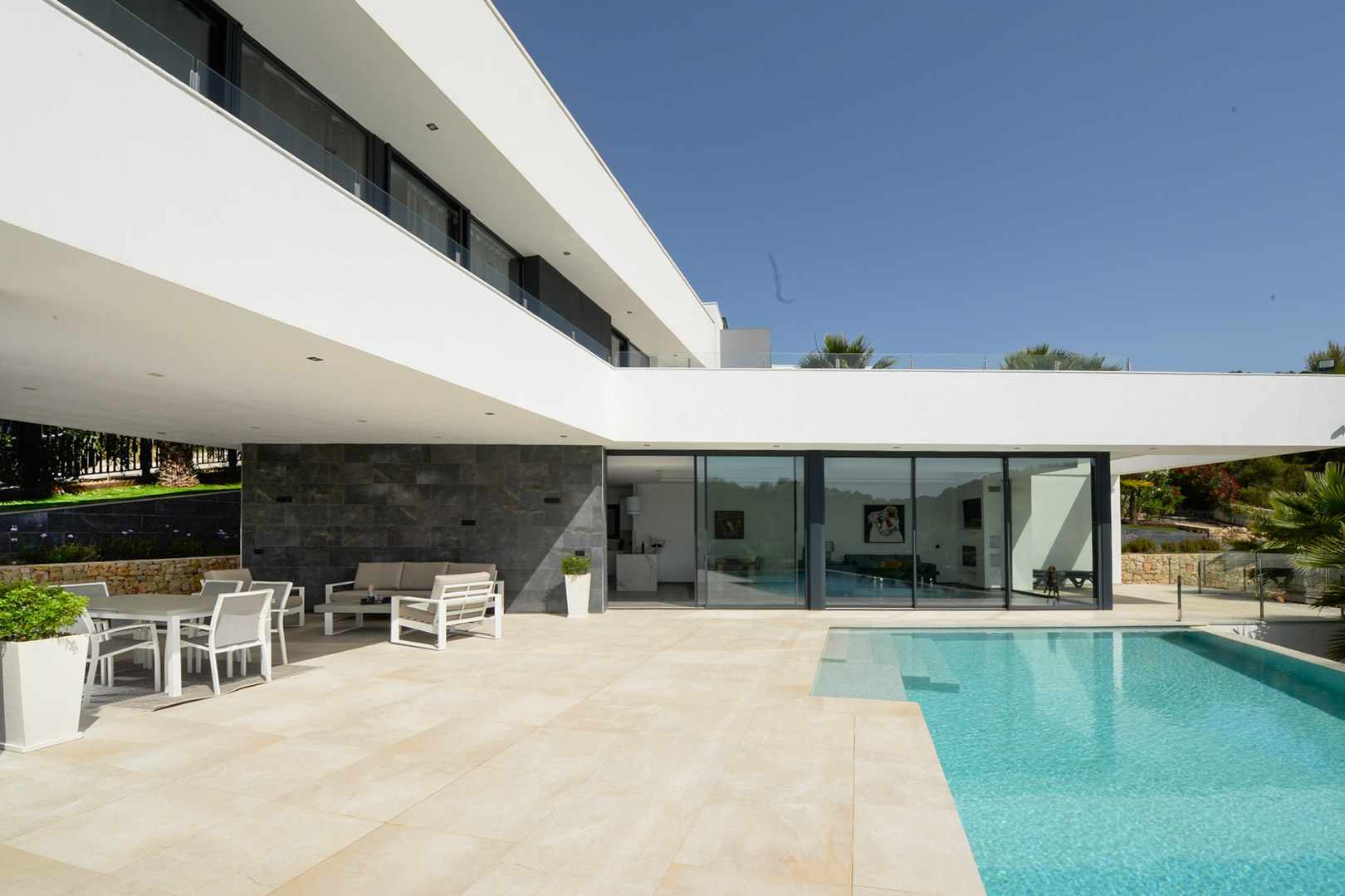 Terraza principal con piscina infinity, zona de comedor exterior bajo porche y fachada minimalista con ventanales de suelo a techo y revestimiento de piedra.
