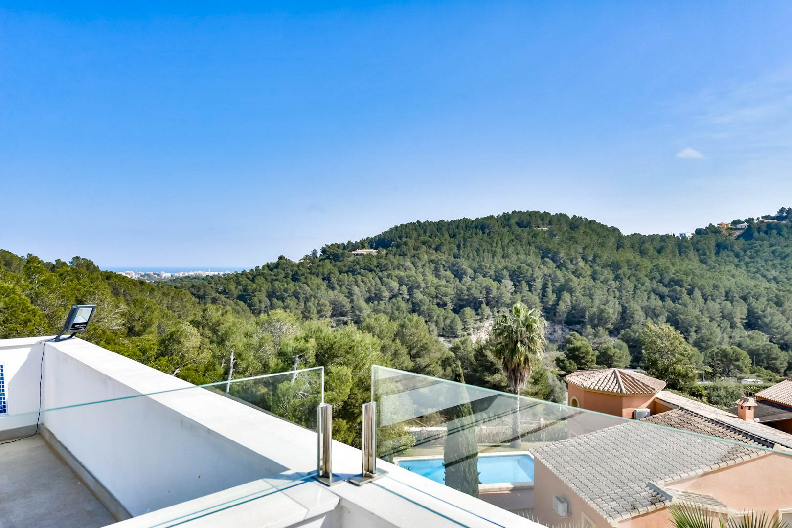 Terraza superior con barandilla de cristal y acero inoxidable, ofreciendo vistas despejadas a las colinas de pinos y al mar Mediterráneo en el horizonte.