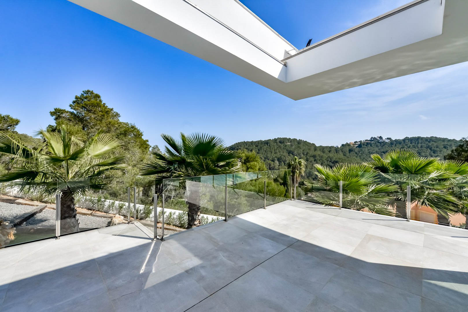 Terraza superior con suelo de gres porcelánico gris, barandillas de cristal y vistas despejadas a las colinas verdes y vegetación mediterránea.