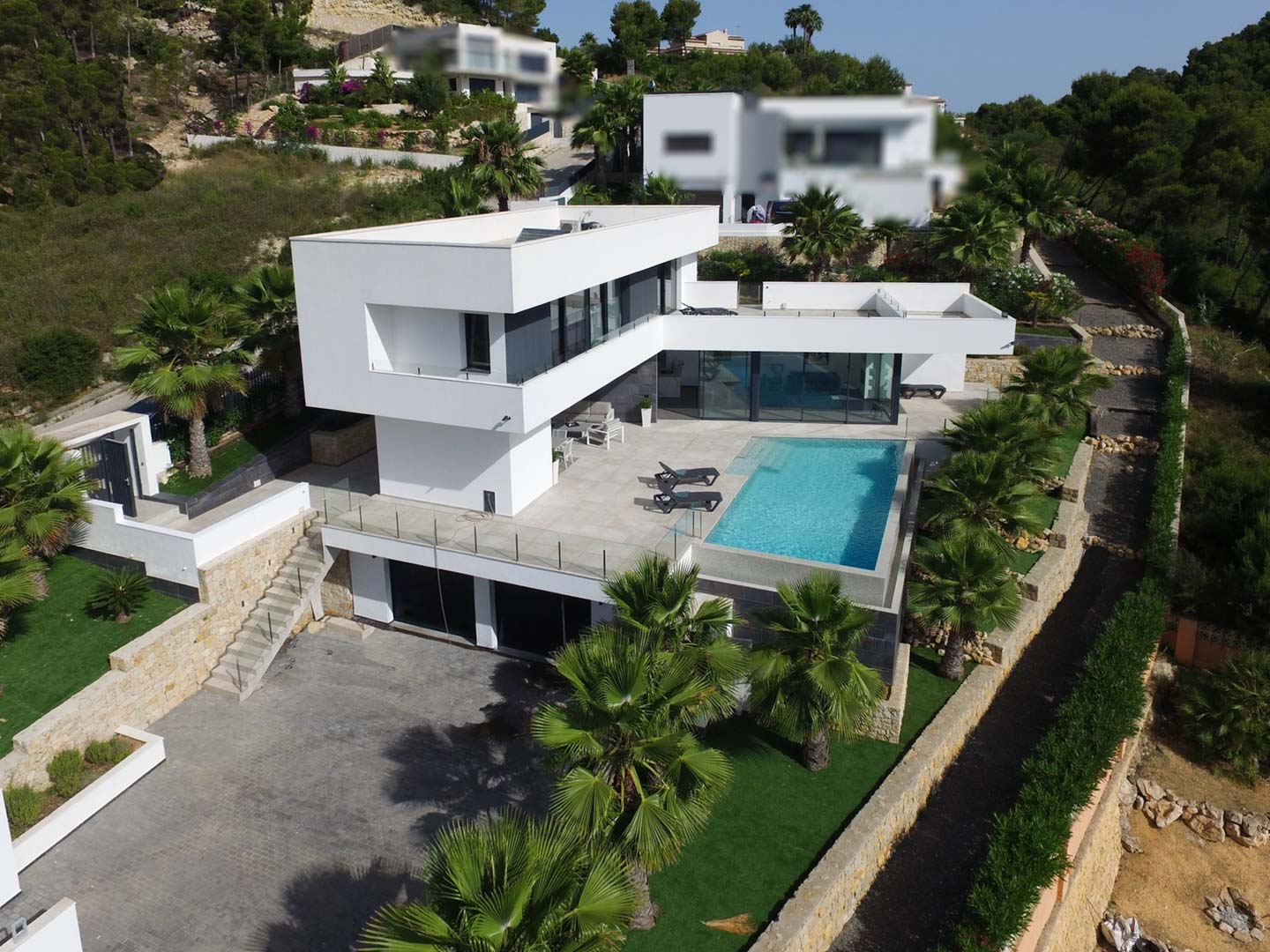 Vista aérea de la villa moderna con arquitectura de líneas blancas, piscina infinity, terrazas de piedra y jardín mediterráneo con palmeras.