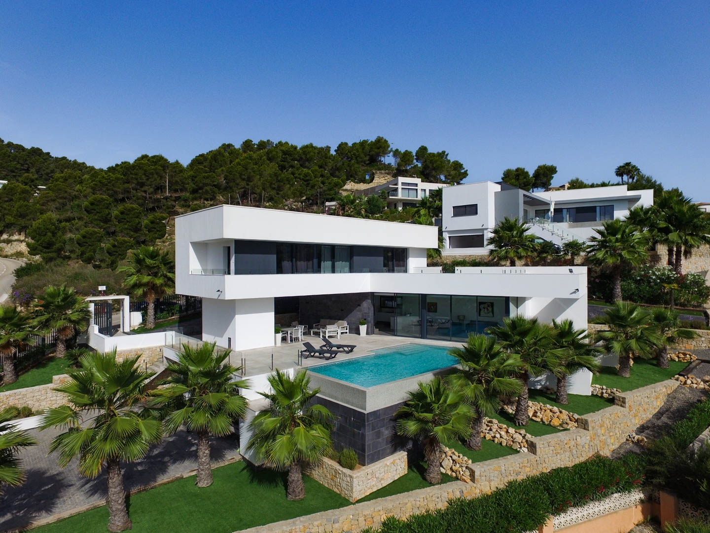 Vista exterior de la villa con arquitectura cúbica blanca, piscina infinity integrada y jardín mediterráneo con palmeras y muros de piedra natural.