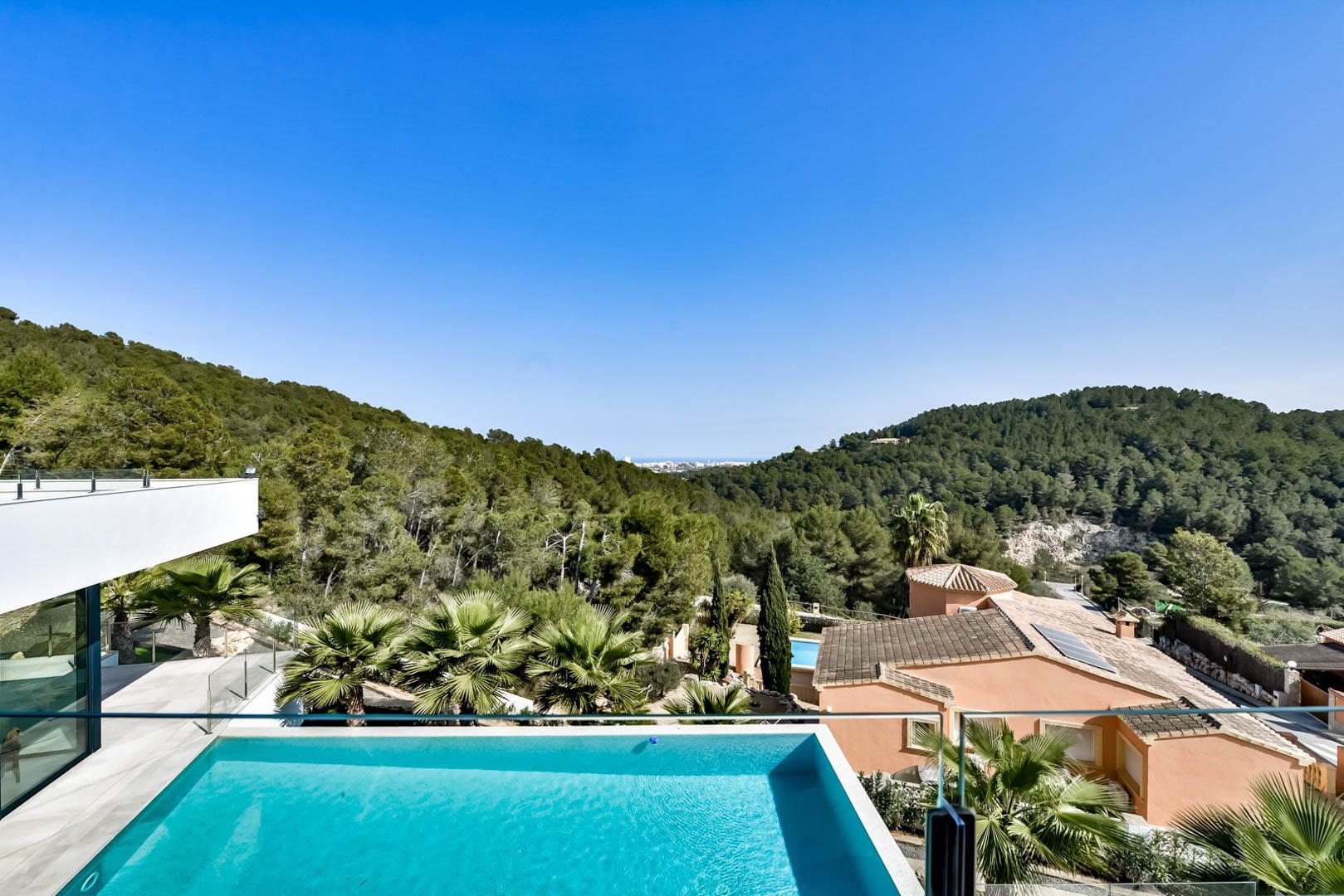 Vista panorámica desde la terraza superior que muestra la piscina infinity, barandillas de cristal y vistas despejadas hacia el mar y las colinas boscosas de Jávea.