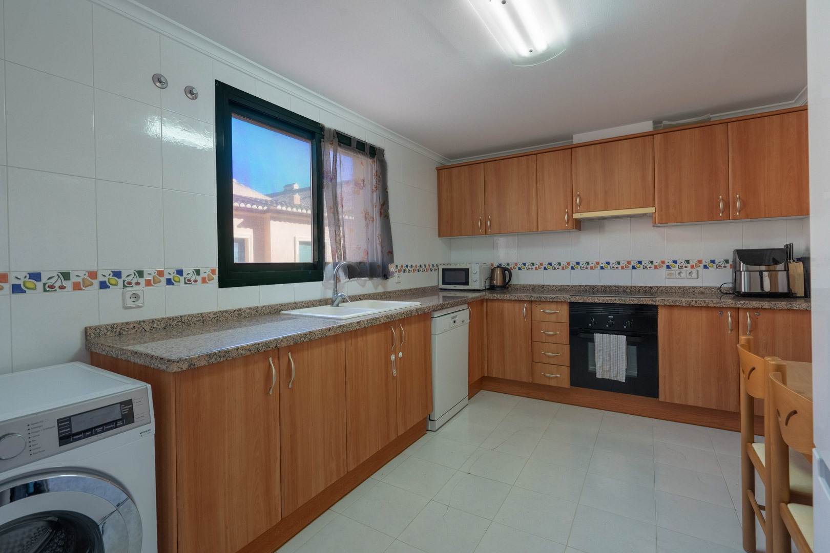 Cocina funcional con mobiliario de madera, encimera de granito, paredes alicatadas con cenefa decorativa y equipamiento completo que incluye lavadora y lavavajillas.