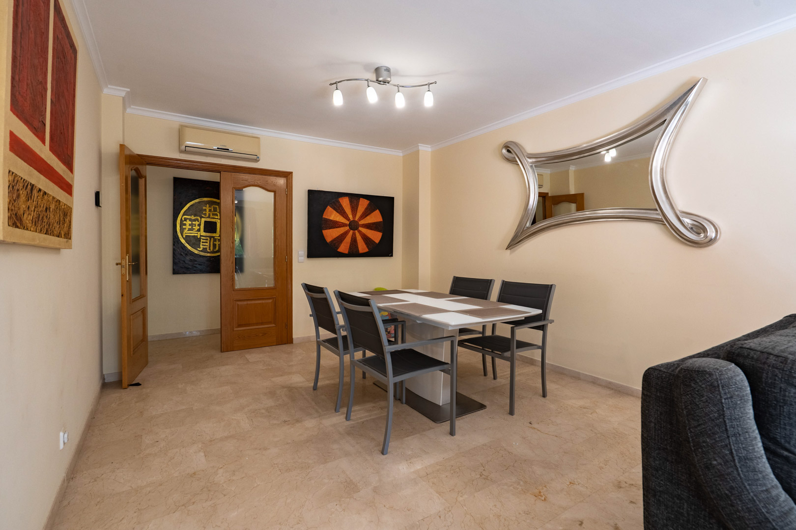 Comedor con suelos de mármol, mesa moderna para cuatro y un gran espejo decorativo, comunicado mediante puertas de madera con el resto de la vivienda.