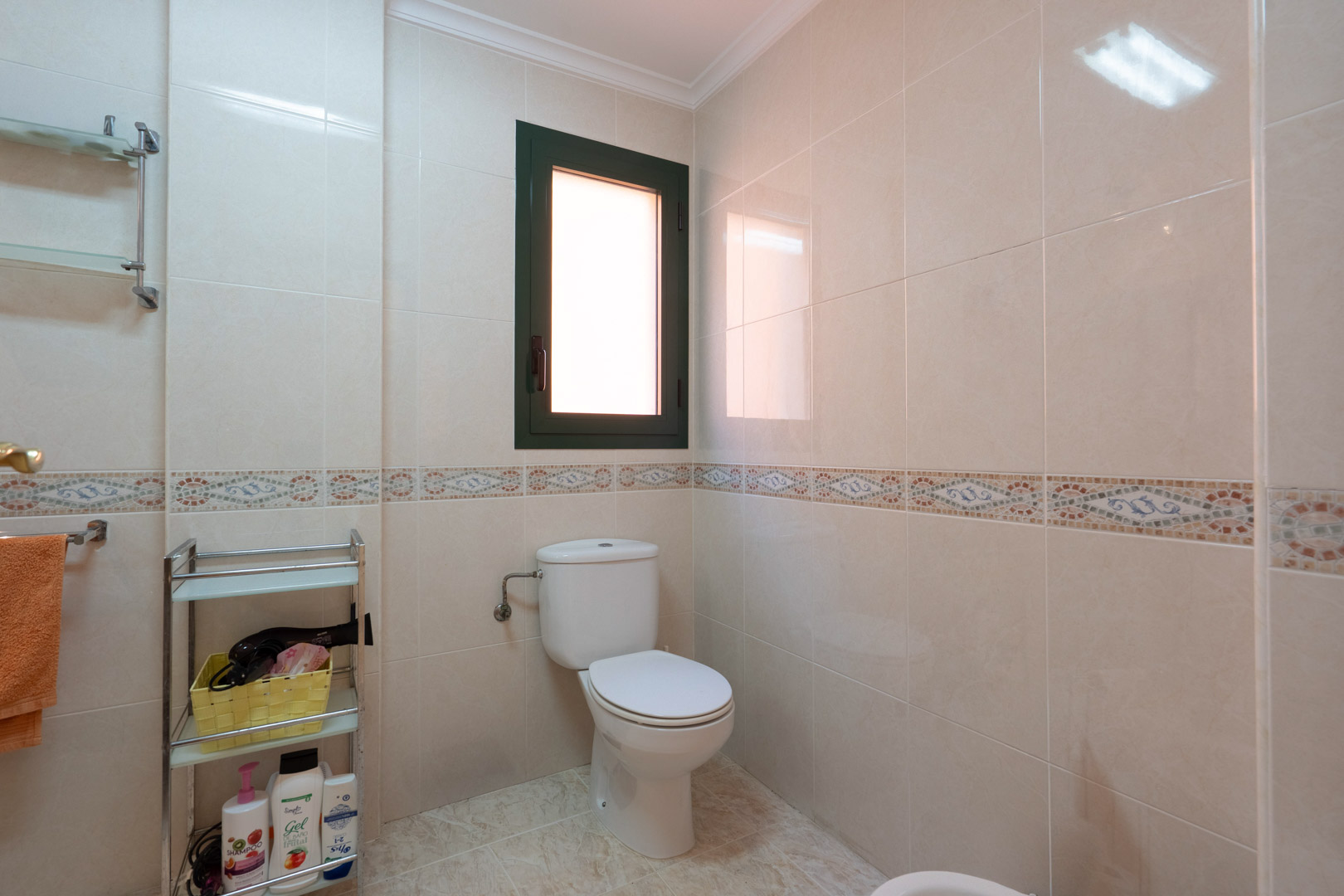 Cuarto de baño con azulejos en tonos crema, cenefa decorativa de mosaico, inodoro blanco y una ventana oscilobatiente que aporta luz natural y ventilación.