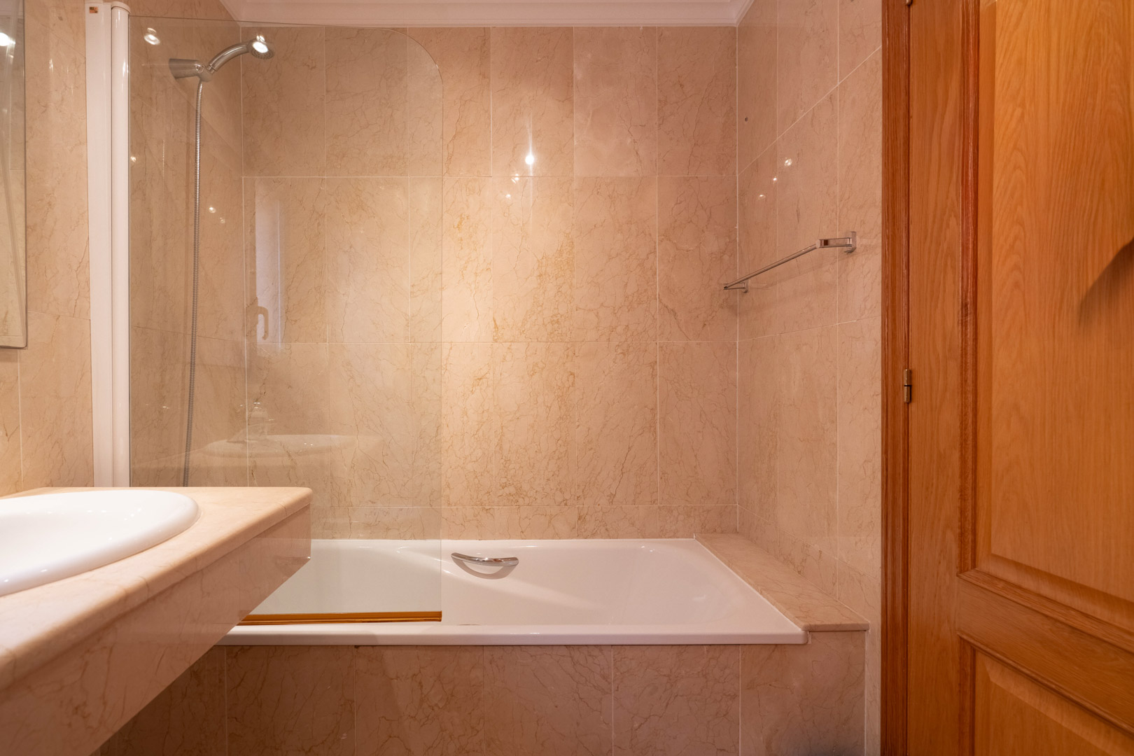 Cuarto de baño con bañera, mampara de cristal y paredes revestidas en mármol crema. Incluye encimera de piedra natural y carpintería de madera de calidad.