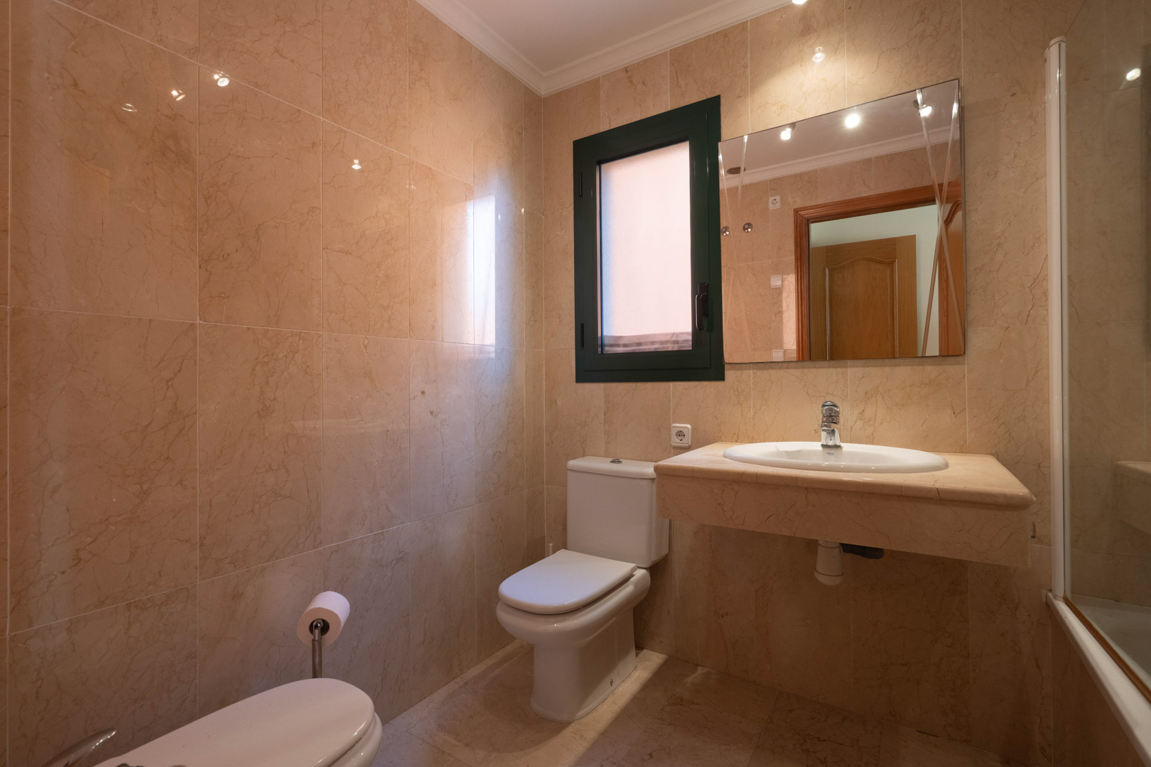 Cuarto de baño revestido íntegramente en mármol crema, con lavabo encastrado en encimera de piedra, sanitarios blancos y ventana para ventilación natural.