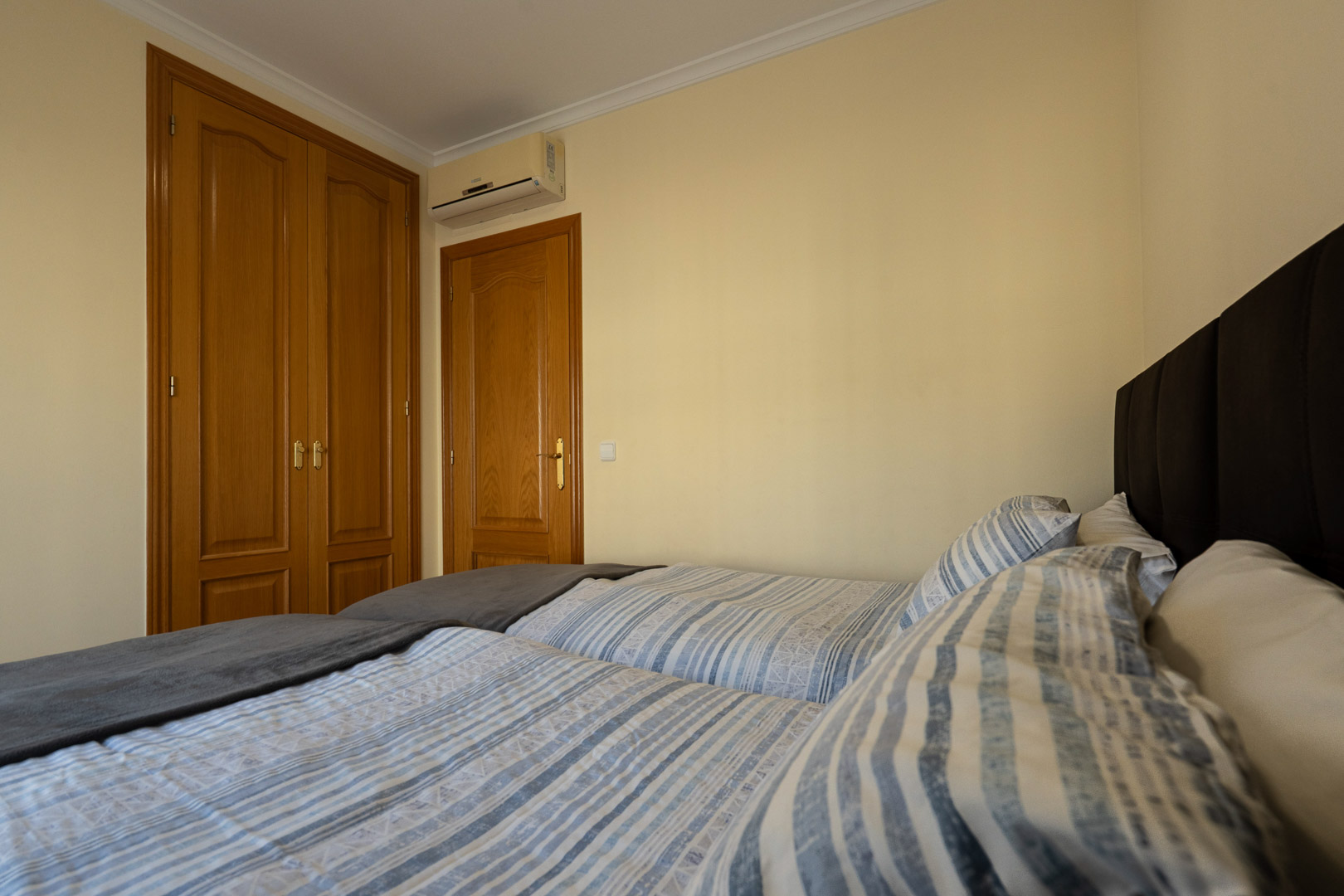 Dormitorio con armarios empotrados de madera de diseño clásico, aire acondicionado individual y camas con cabecero tapizado en tonos oscuros.