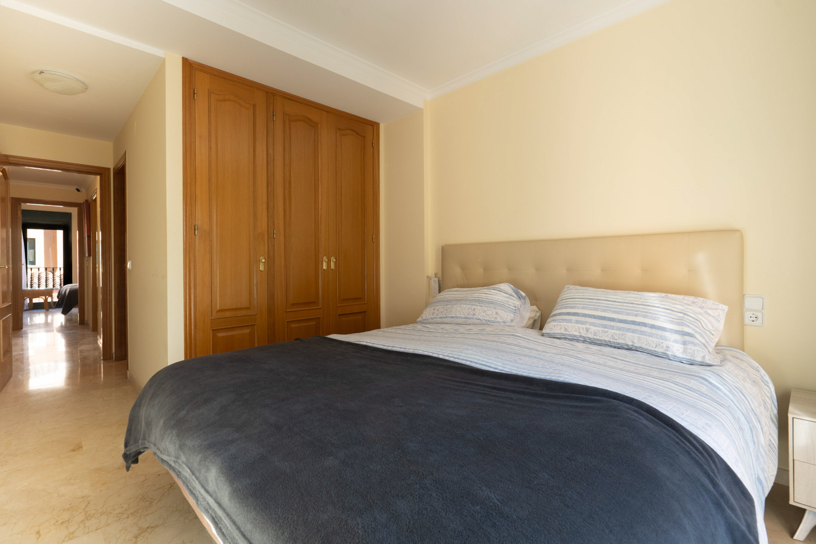 Dormitorio con armarios empotrados de madera, suelos de mármol y un cabecero tapizado en tonos neutros, conectado al pasillo principal de la vivienda.
