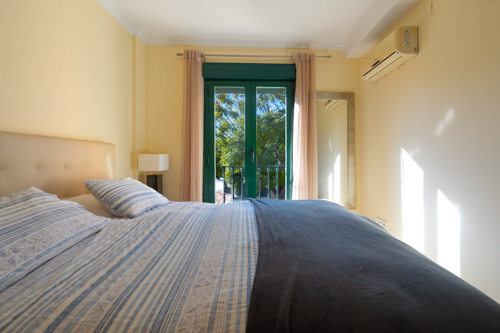 Dormitorio con cama doble, cabecero acolchado y aire acondicionado. Las puertas verdes dan acceso a un balcón francés con vistas a los árboles.