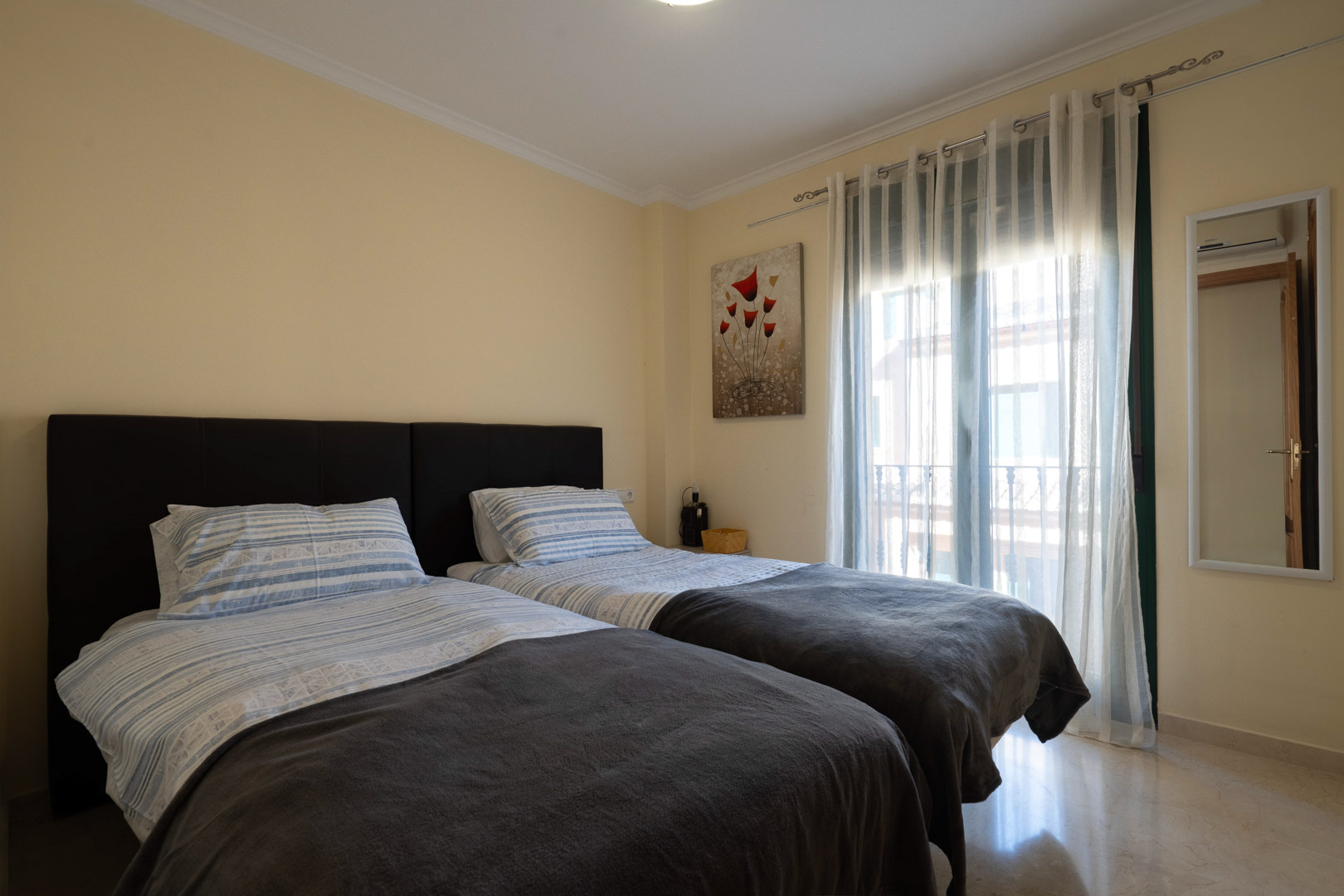 Dormitorio luminoso con dos camas individuales, suelos de mármol pulido y acceso directo al balcón a través de ventanales con cortinas ligeras.