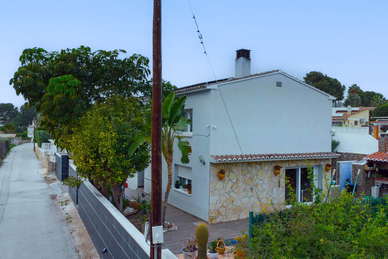 Vista exterior de la villa con fachada blanca y revestimiento de piedra natural, rodeada de vegetación mediterránea y equipada con paneles solares.
