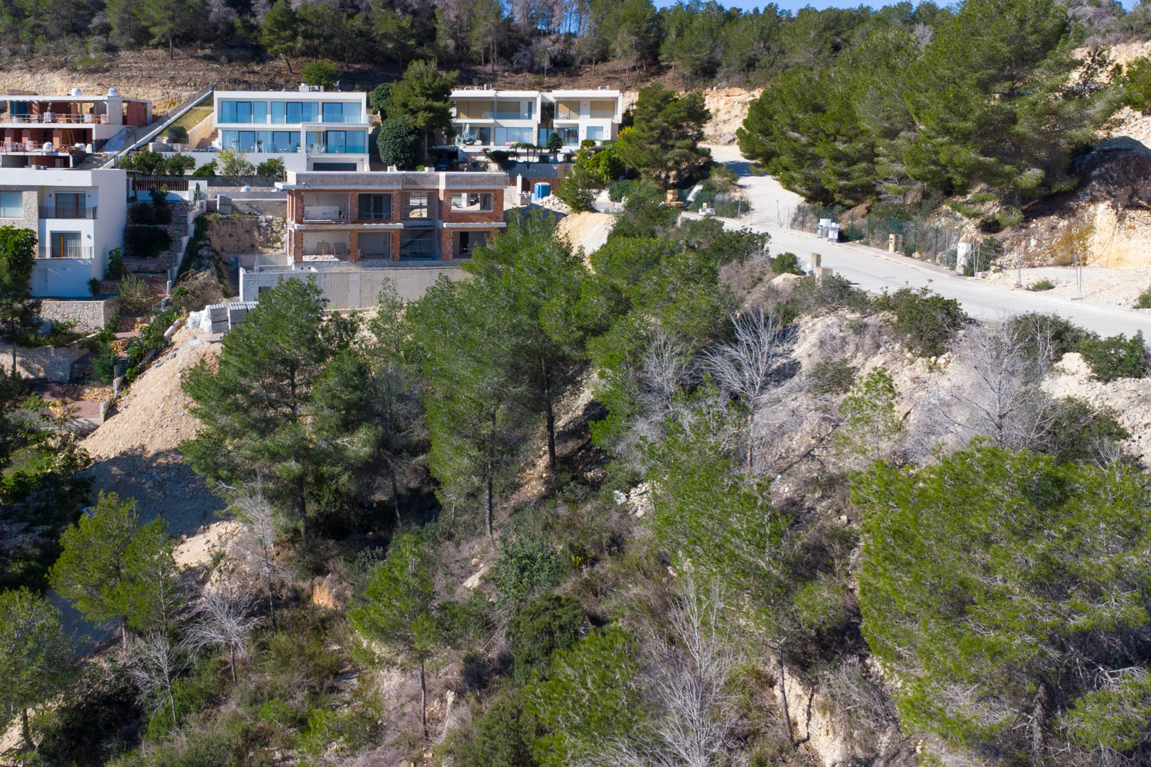 Parcela elevada en la urbanización Tosalet 5, rodeada de pinos y villas modernas. Se observa el acceso asfaltado y una construcción en curso sobre el terreno inclinado.