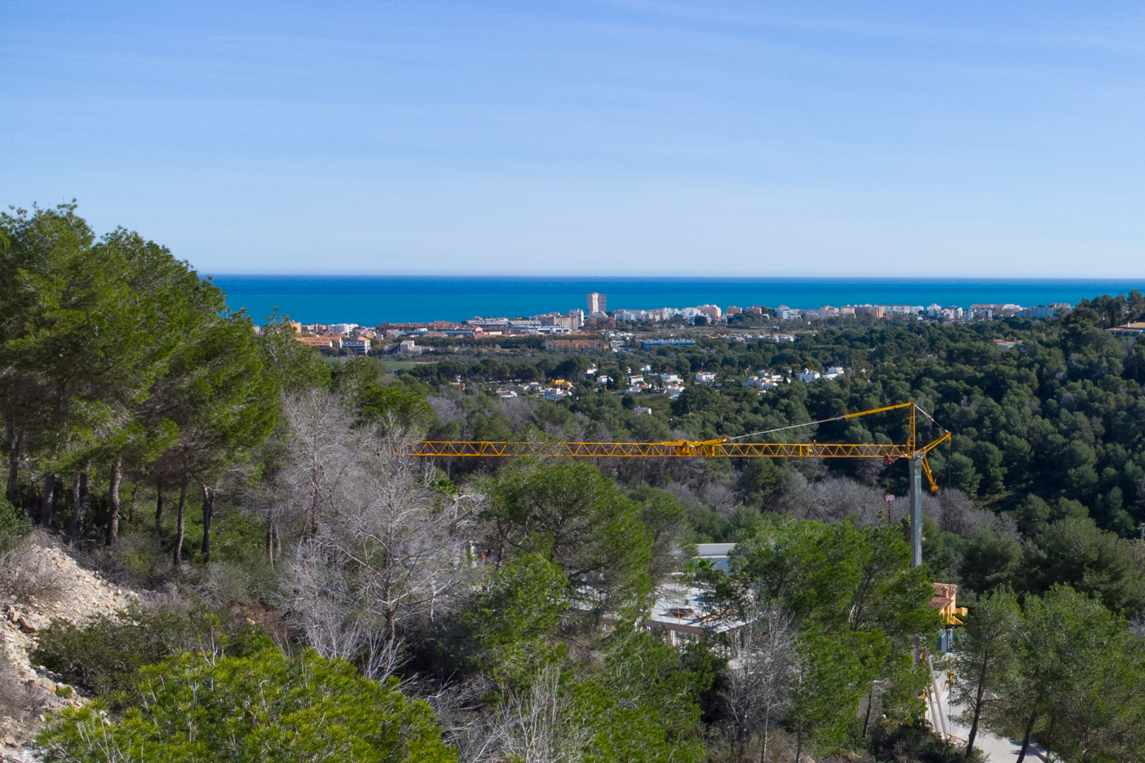 Vista panorámica al mar Mediterráneo desde la parcela elevada, rodeada de pinos autóctonos y con el núcleo urbano de Jávea al fondo bajo un cielo despejado.