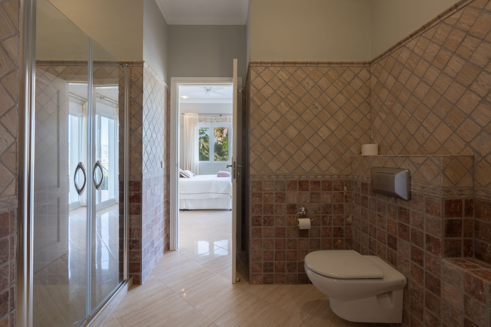 Baño con azulejos rústicos en tonos tierra, inodoro suspendido y acceso directo al dormitorio principal inundado de luz natural.
