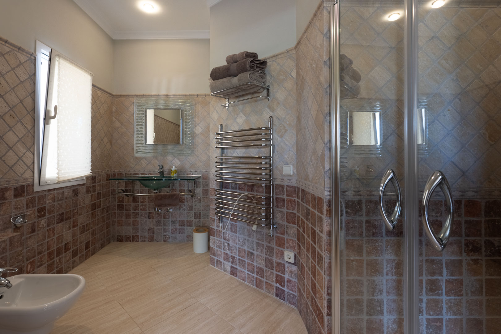 Baño con revestimiento cerámico en tonos tierra, lavabo de cristal suspendido, radiador toallero cromado y cabina de ducha con mampara de vidrio.