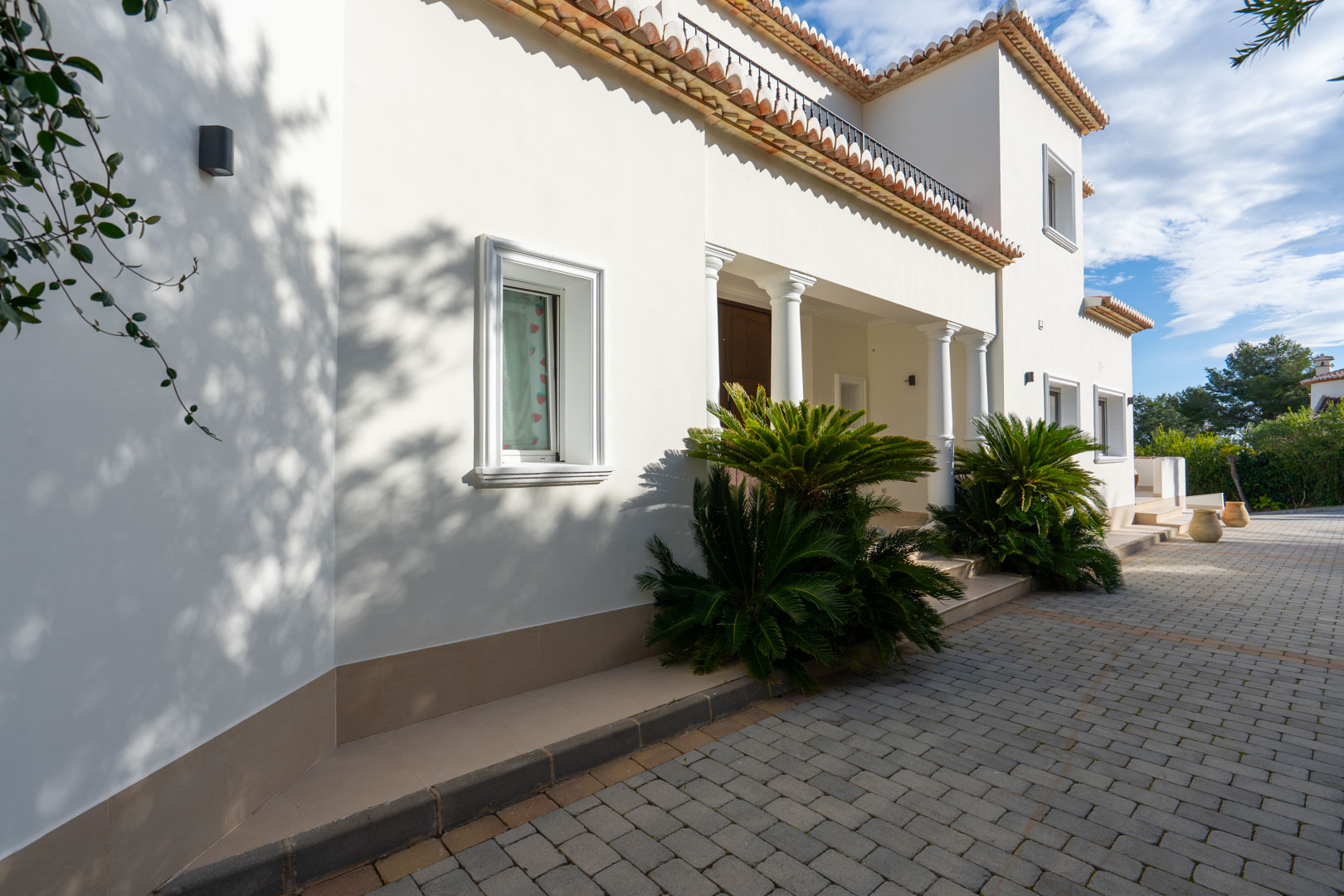 Fachada principal de estilo mediterráneo con columnas blancas, entrada pavimentada y detalles de vegetación junto a la puerta de acceso.