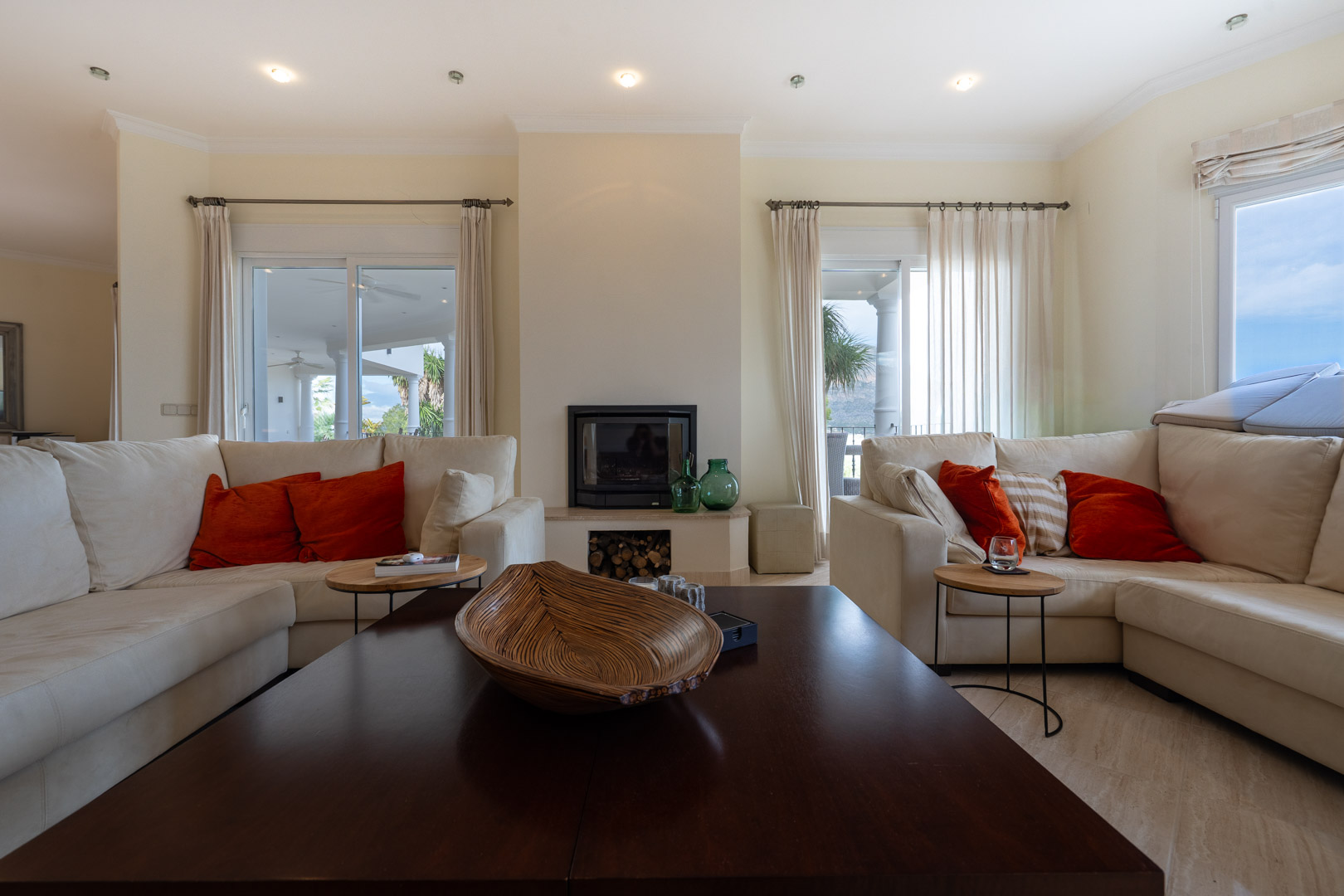 Salón luminoso con chimenea central, sofás en tonos crema y acceso directo a la terraza a través de grandes ventanales con cortinas blancas.