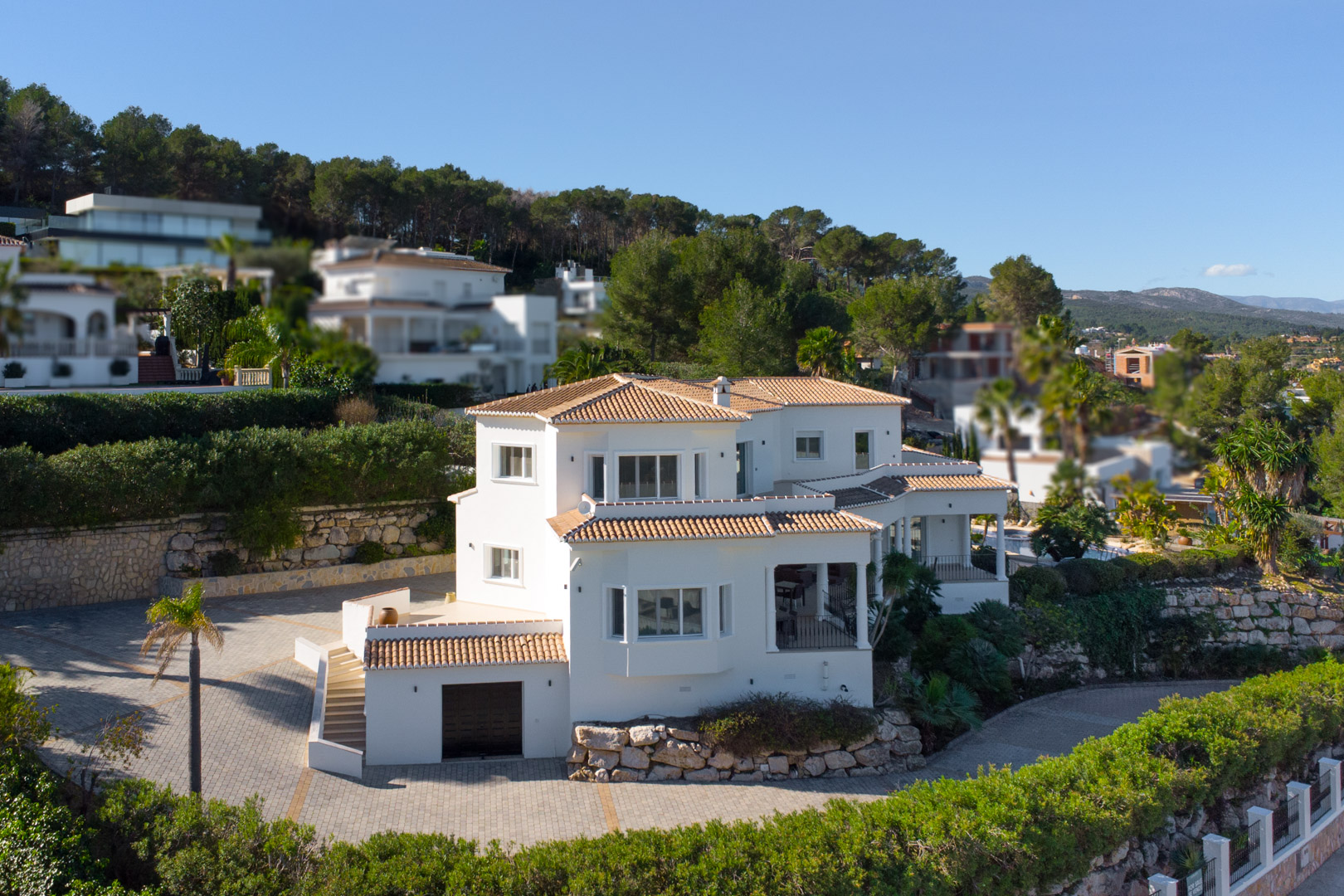 Vista exterior de la villa con fachada blanca y tejados de teja cerámica, destacando el amplio acceso pavimentado y el garaje integrado en la planta inferior.