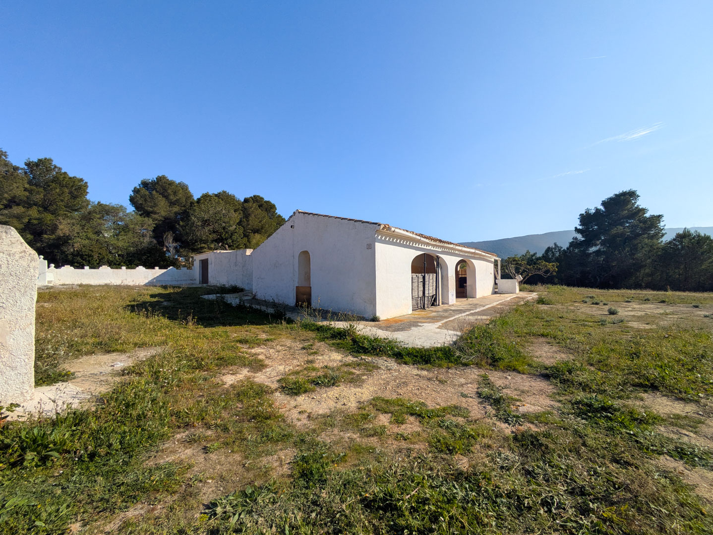 Fachada de la finca tradicional con muros blancos y porches de arco, ubicada en una extensa parcela rústica rodeada de pinos y vegetación autóctona.