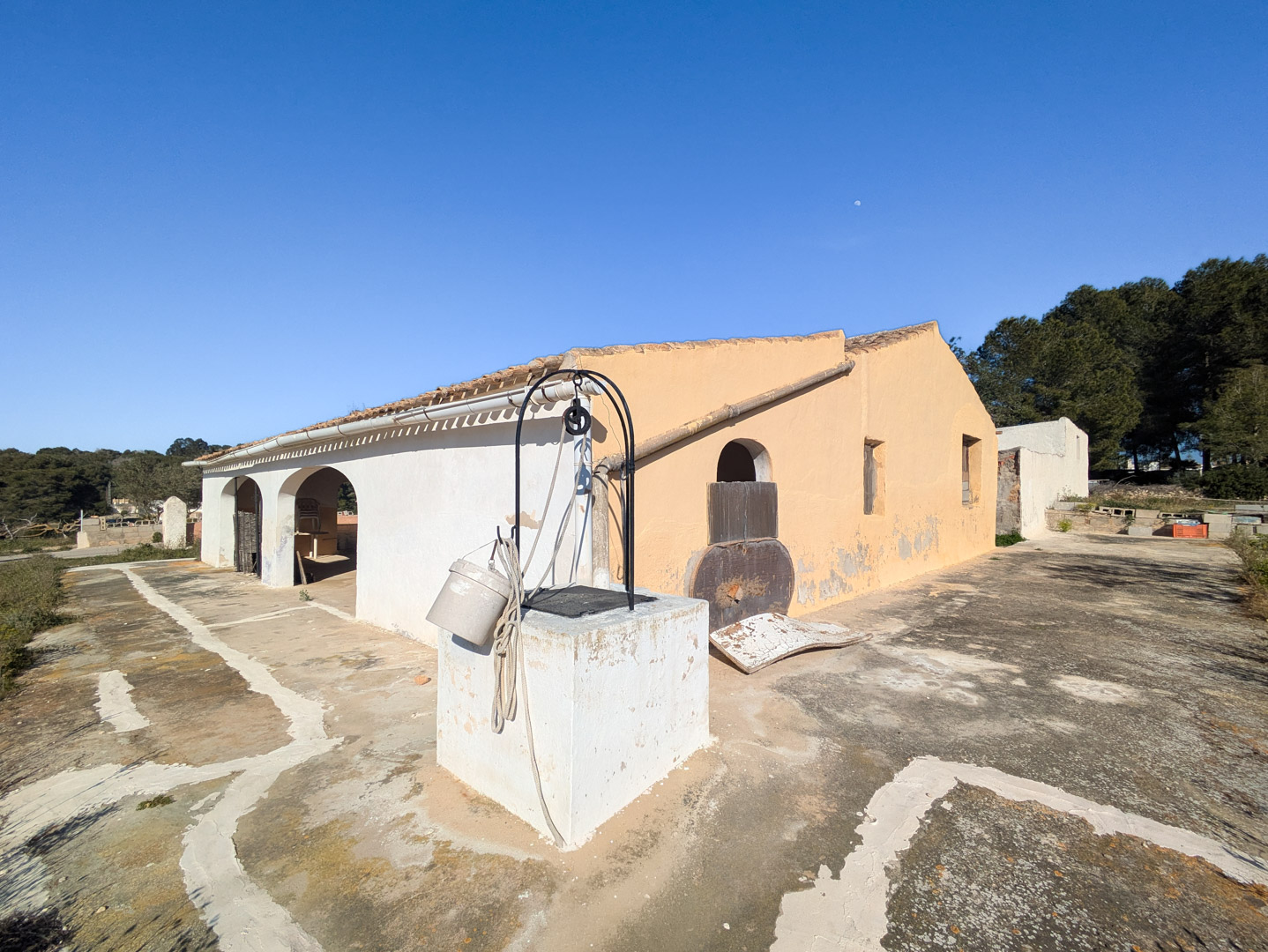 Fachada de la finca tradicional con porche de arcos, pozo antiguo en primer plano y amplia terraza de piedra bajo un cielo despejado.