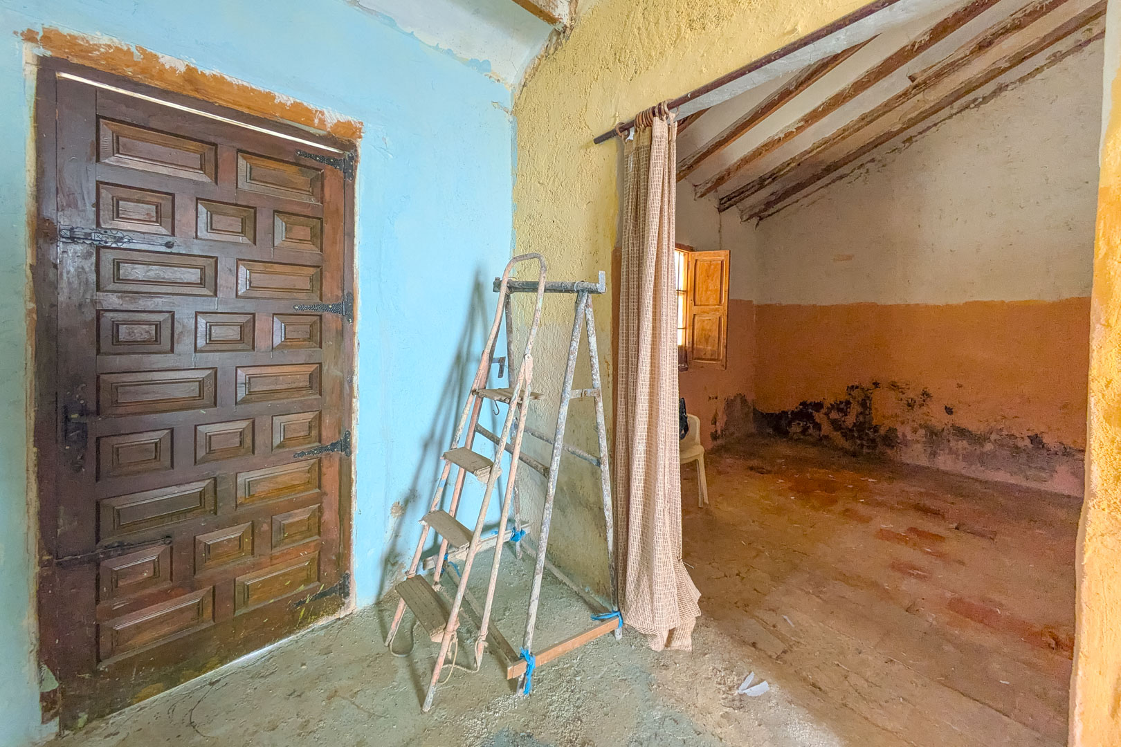 Interior rústico con puerta de madera labrada, paredes de colores y techos con vigas vistas, mostrando el potencial de reforma integral de la finca.