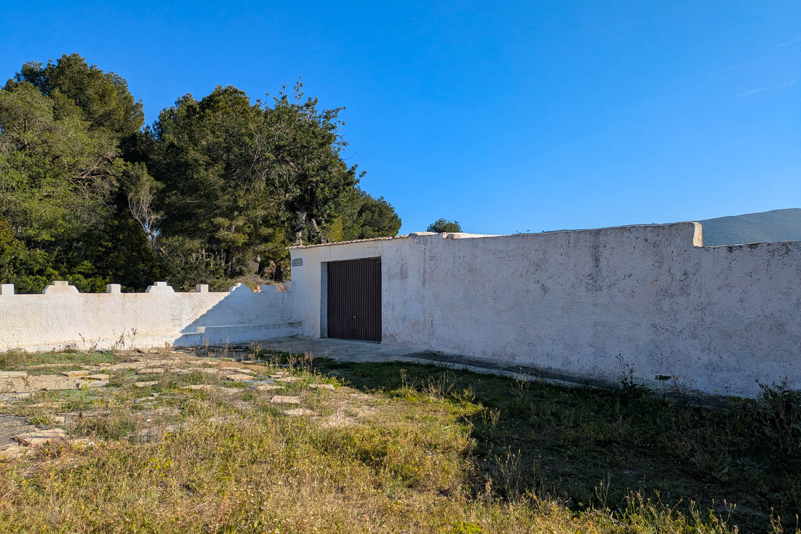 Muro perimetral blanco con puerta de acceso oscura, situado en una parcela rústica rodeada de pinos y vegetación autóctona en Jávea.