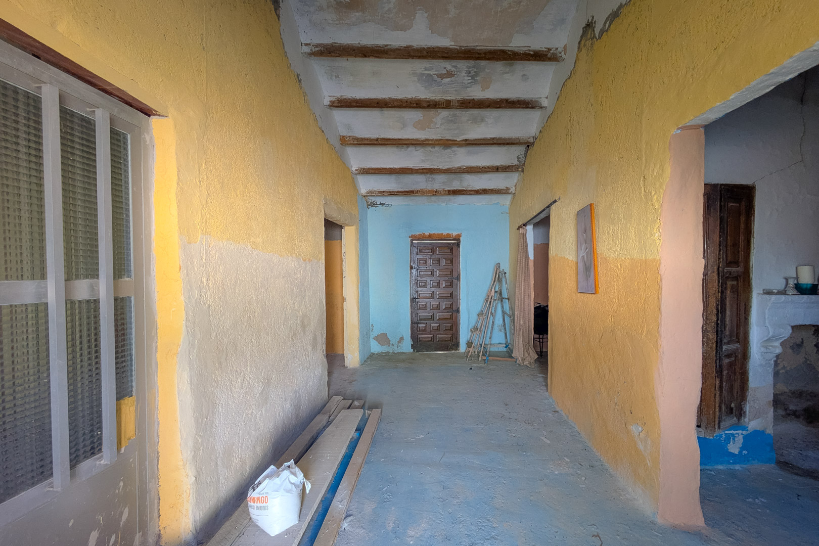 Pasillo interior con techos altos de vigas de madera expuestas y paredes rústicas en tonos amarillos y azules, mostrando el estado original y potencial de reforma.