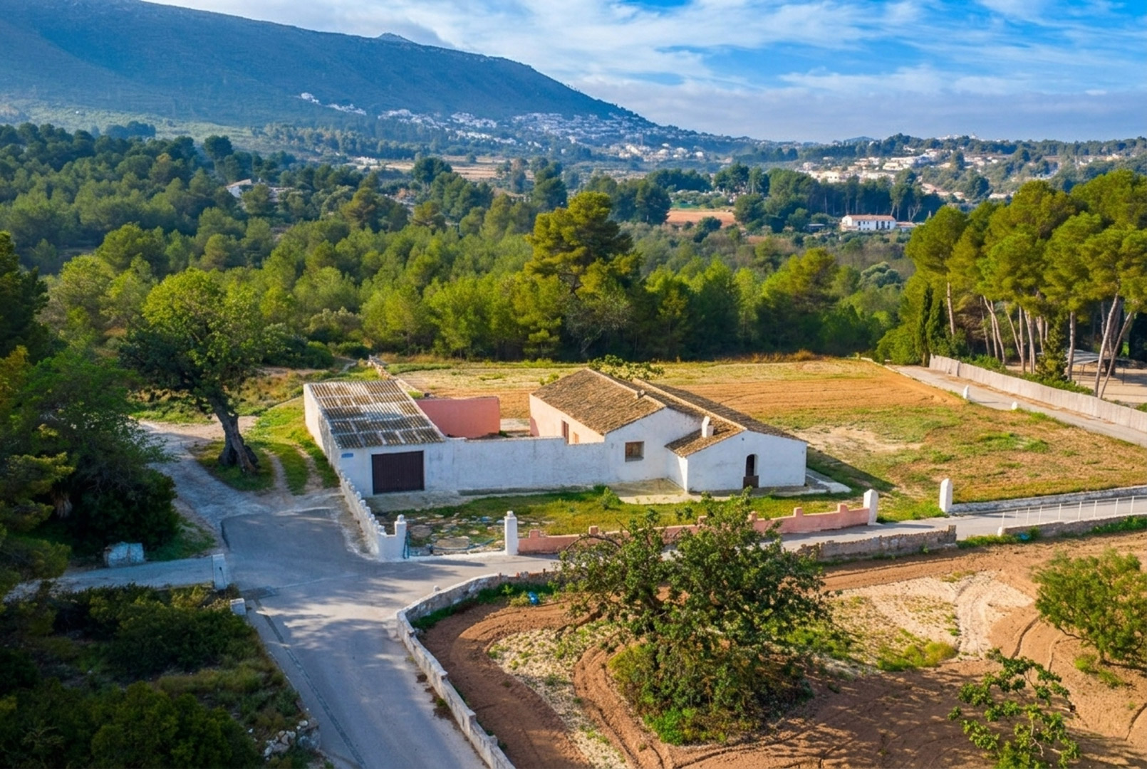 Vista aérea de la finca tradicional con muros blancos y tejados de teja, situada en una amplia parcela llana rodeada de frondosos bosques de pinos y montañas.