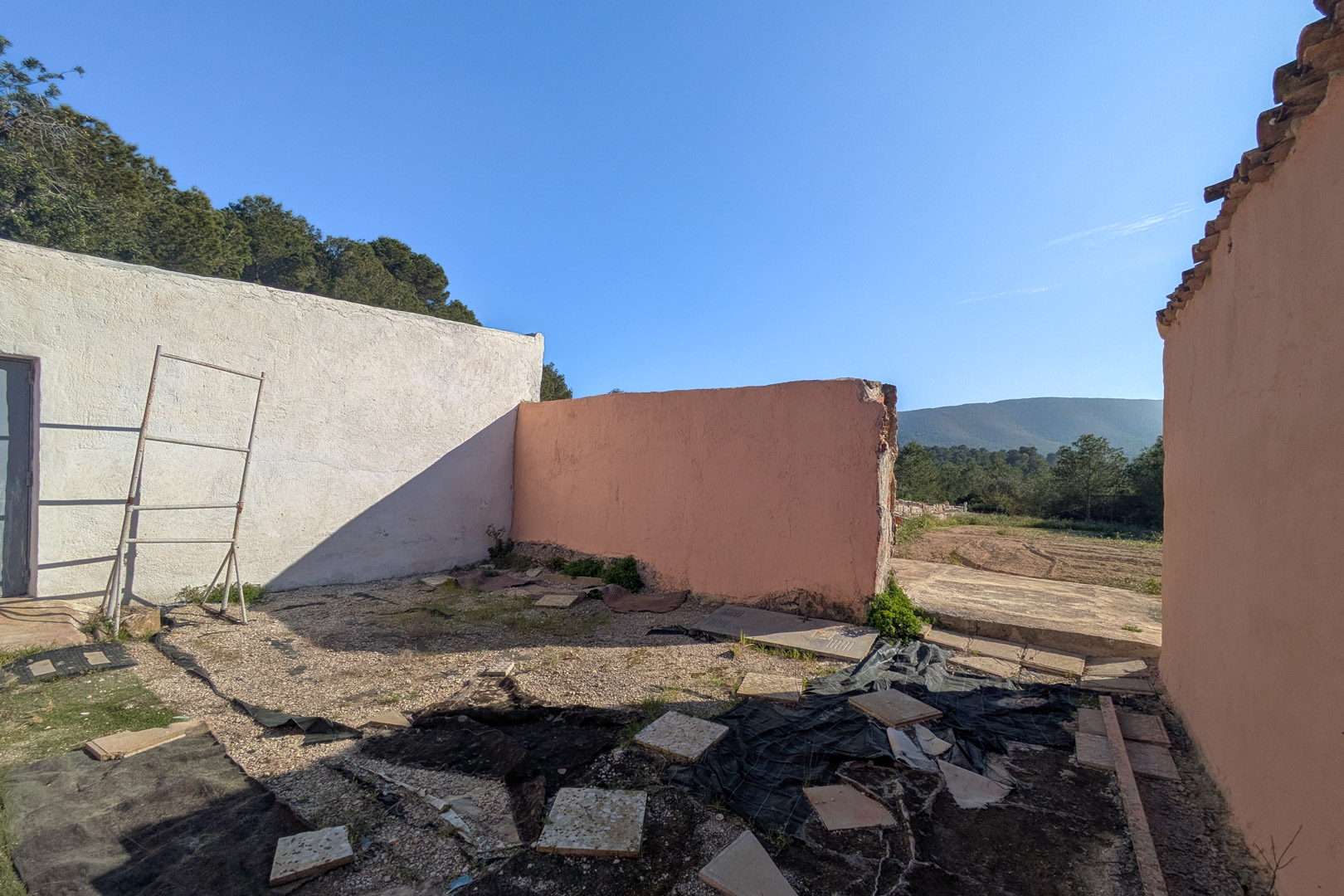 Vista de una zona exterior de la finca con muros de revoco en blanco y terracota, mostrando el estado actual de la estructura y las vistas despejadas hacia las montañas.